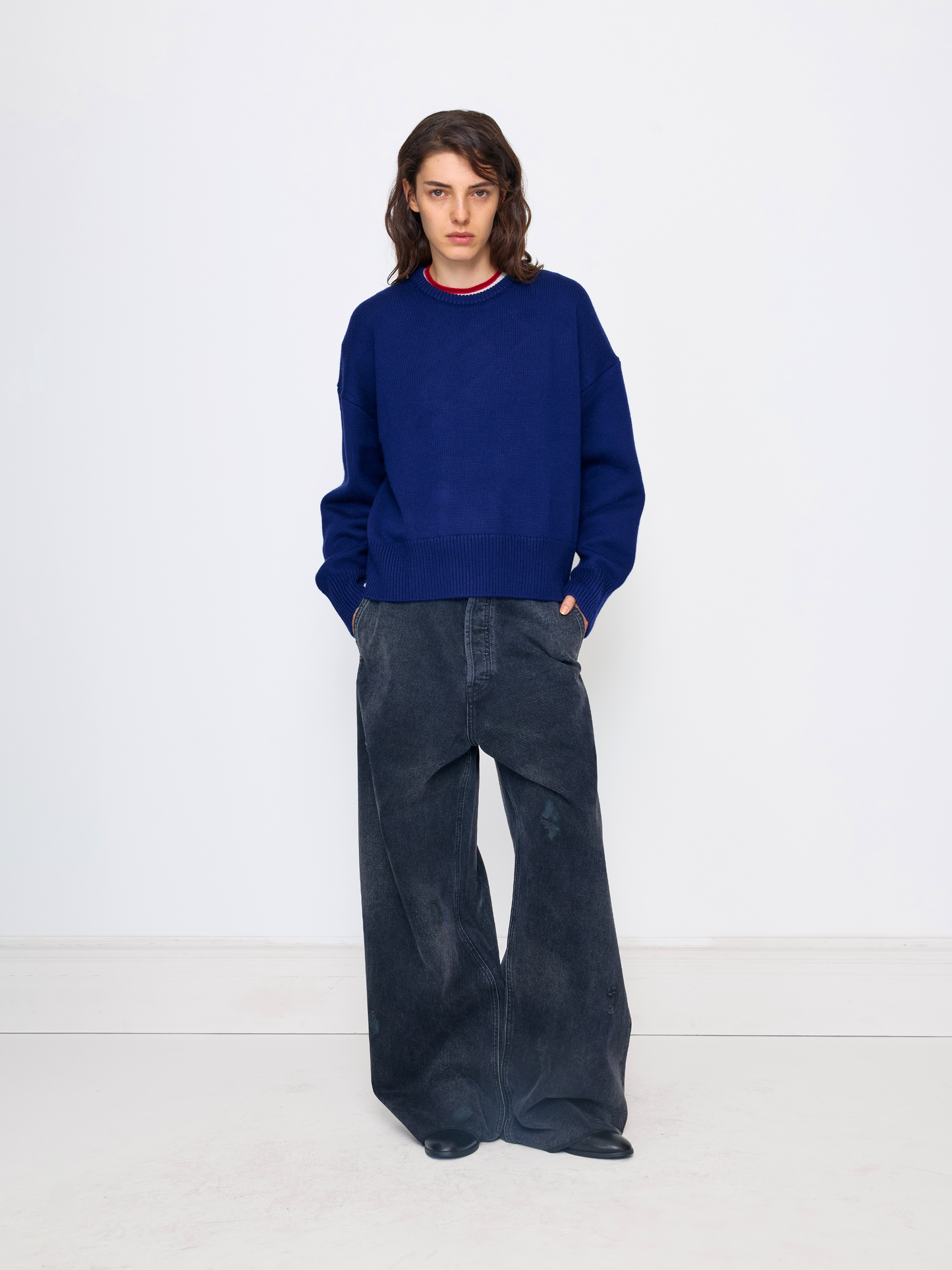A model stands against a plain white studio wall wearing a navy blue crewneck sweater with a thin red and white striped collar, paired with dark wide-leg corduroy pants.