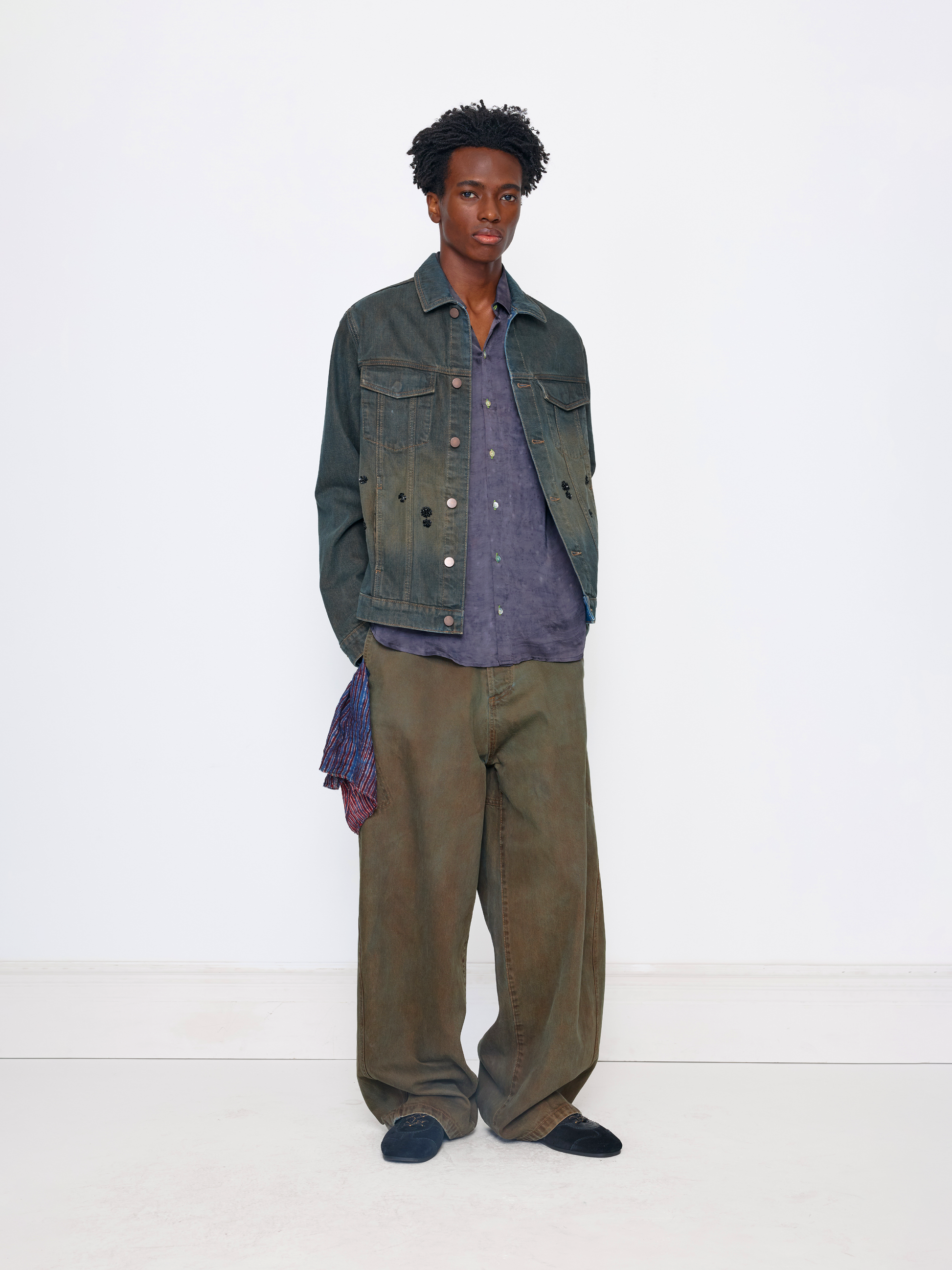 A model stands against a plain white studio background wearing a dark green distressed denim jacket over a purple button-up shirt, paired with wide-leg brownish-green trousers and dark slip-on shoes.