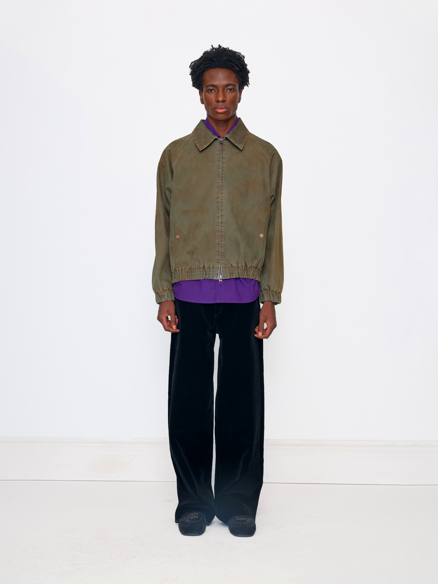 A model stands against a plain white studio background wearing an olive green cropped jacket over a purple shirt, paired with black velvet trousers and dark loafers.