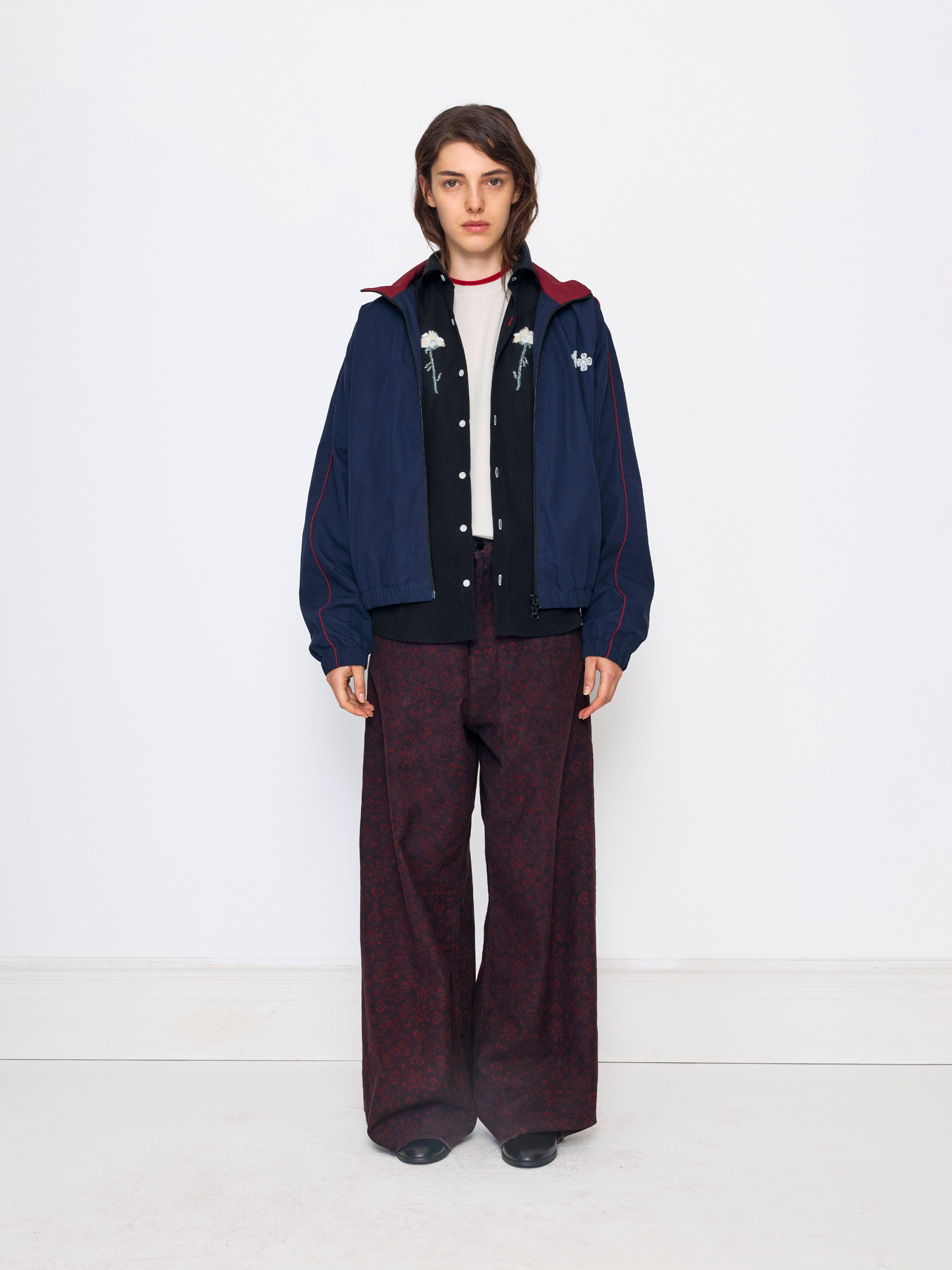 A model stands against a plain white studio background wearing a dark navy jacket with red lining and white embroidered details, a black button-front cardigan, a white top, and dark patterned wide-leg trousers.