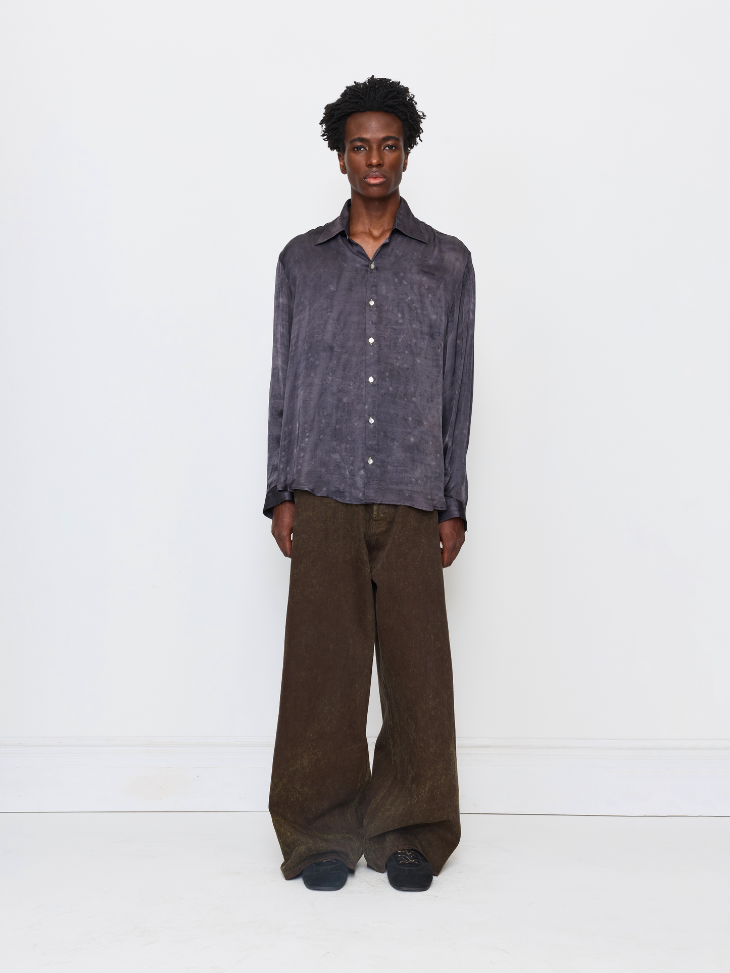 A model stands against a plain white studio background wearing a dark gray, textured long-sleeve button-up shirt and wide-leg brown trousers.