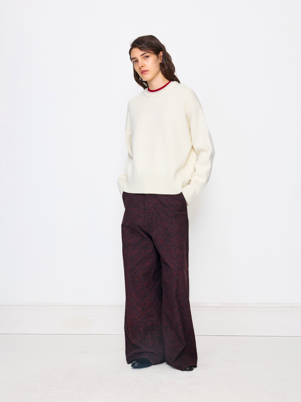 A model stands against a clean white studio background wearing a cream knit mock-neck sweater and dark burgundy wide-leg trousers with a subtle damask pattern.