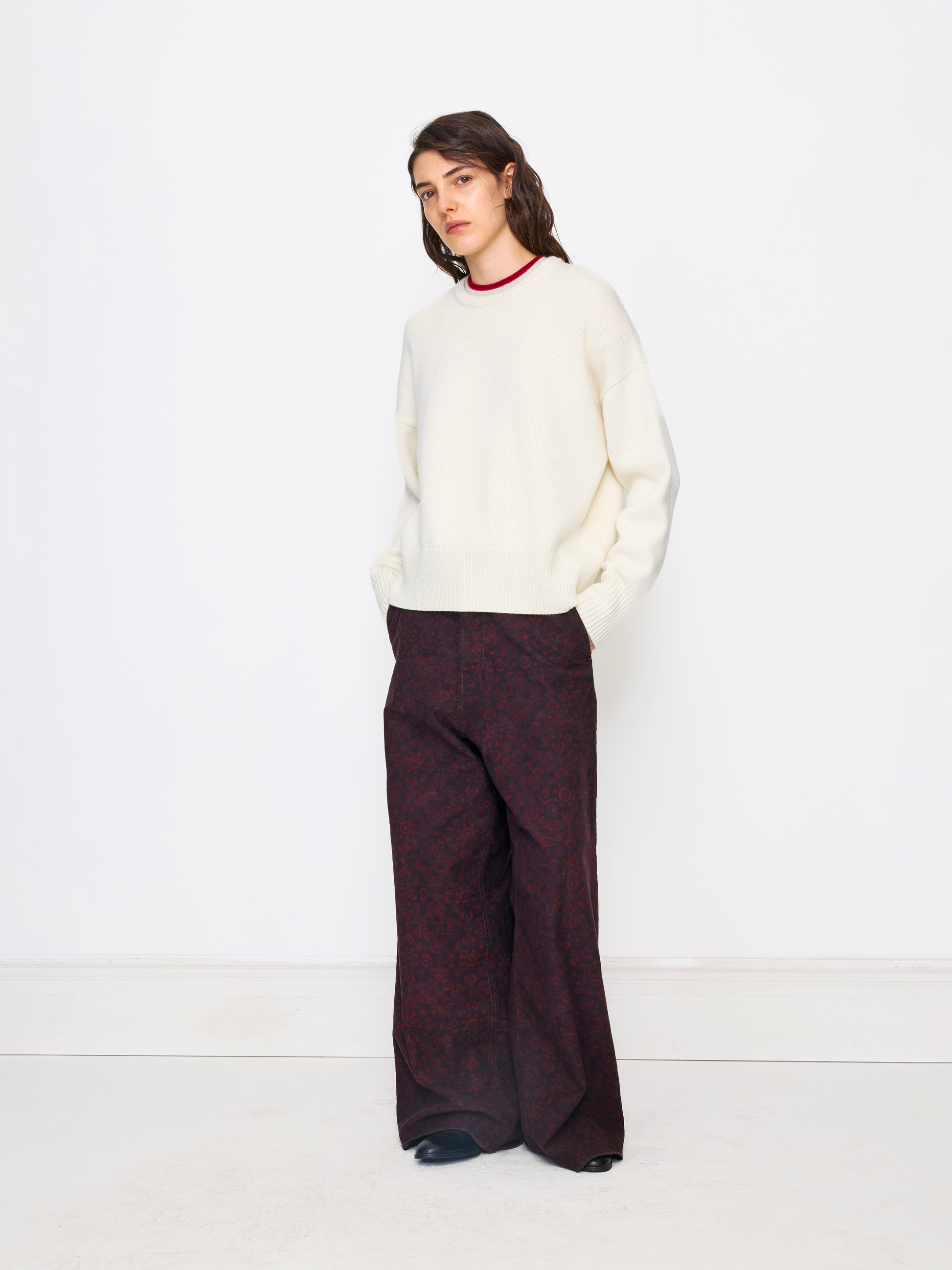 A model stands against a clean white studio background wearing a cream knit mock-neck sweater and dark burgundy wide-leg trousers with a subtle damask pattern.
