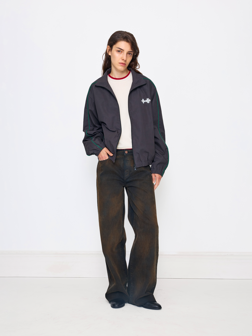 A model stands against a plain white studio background wearing a dark navy zip-up jacket with green stripes on the sleeves, a white crewneck shirt, dark brown wide-leg jeans, and black shoes.