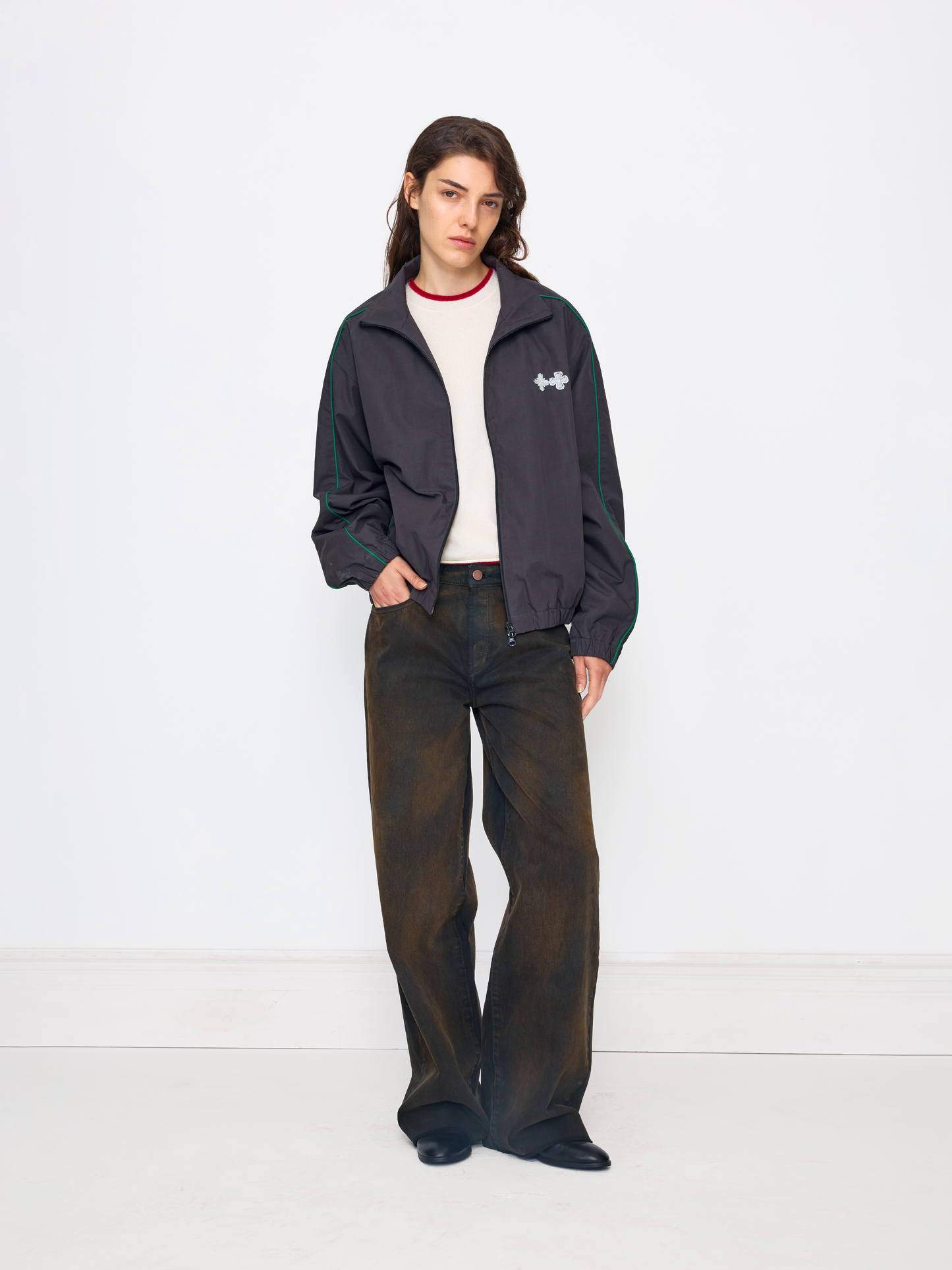 A model stands against a plain white studio background wearing a dark navy zip-up jacket with green stripes on the sleeves, a white crewneck shirt, dark brown wide-leg jeans, and black shoes.