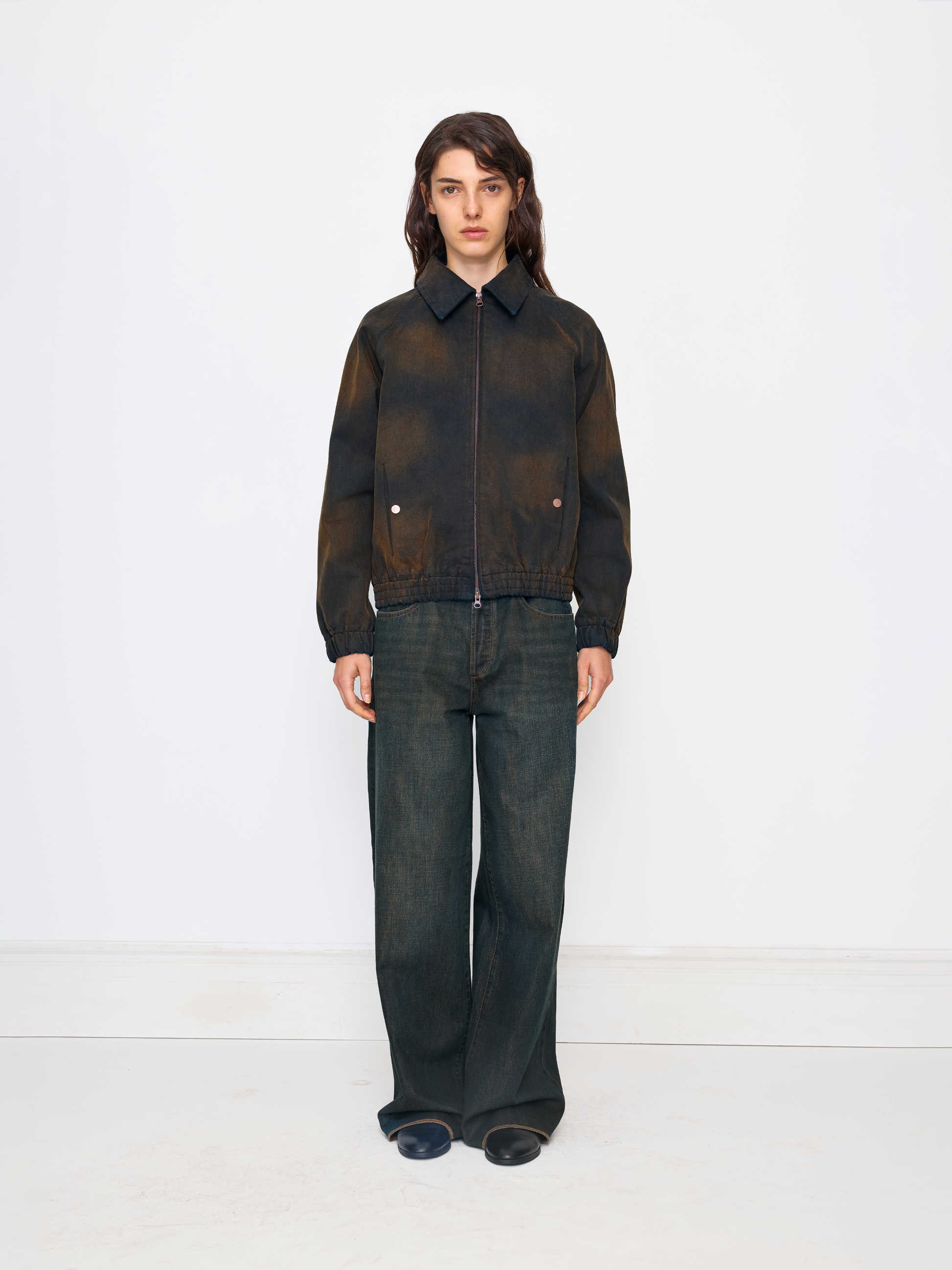 A model stands against a plain white studio background wearing a dark brown and black tie-dye zip-up jacket with dark wide-leg jeans.