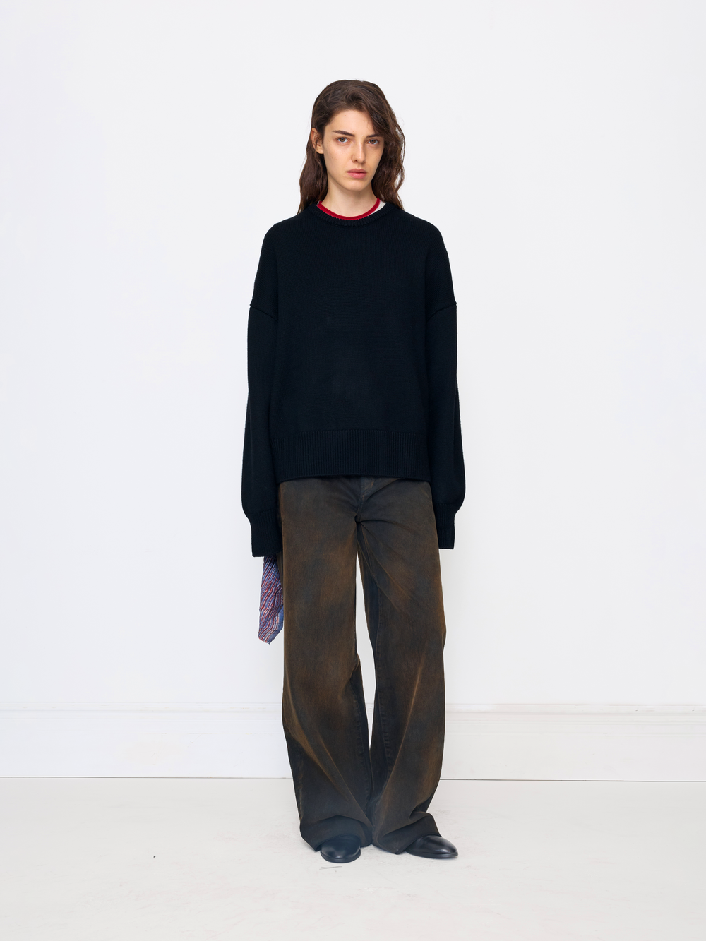 A model stands against a plain white studio wall wearing an oversized black knit sweater with a thin red collar trim, loose dark brown wide-leg trousers, and black shoes.