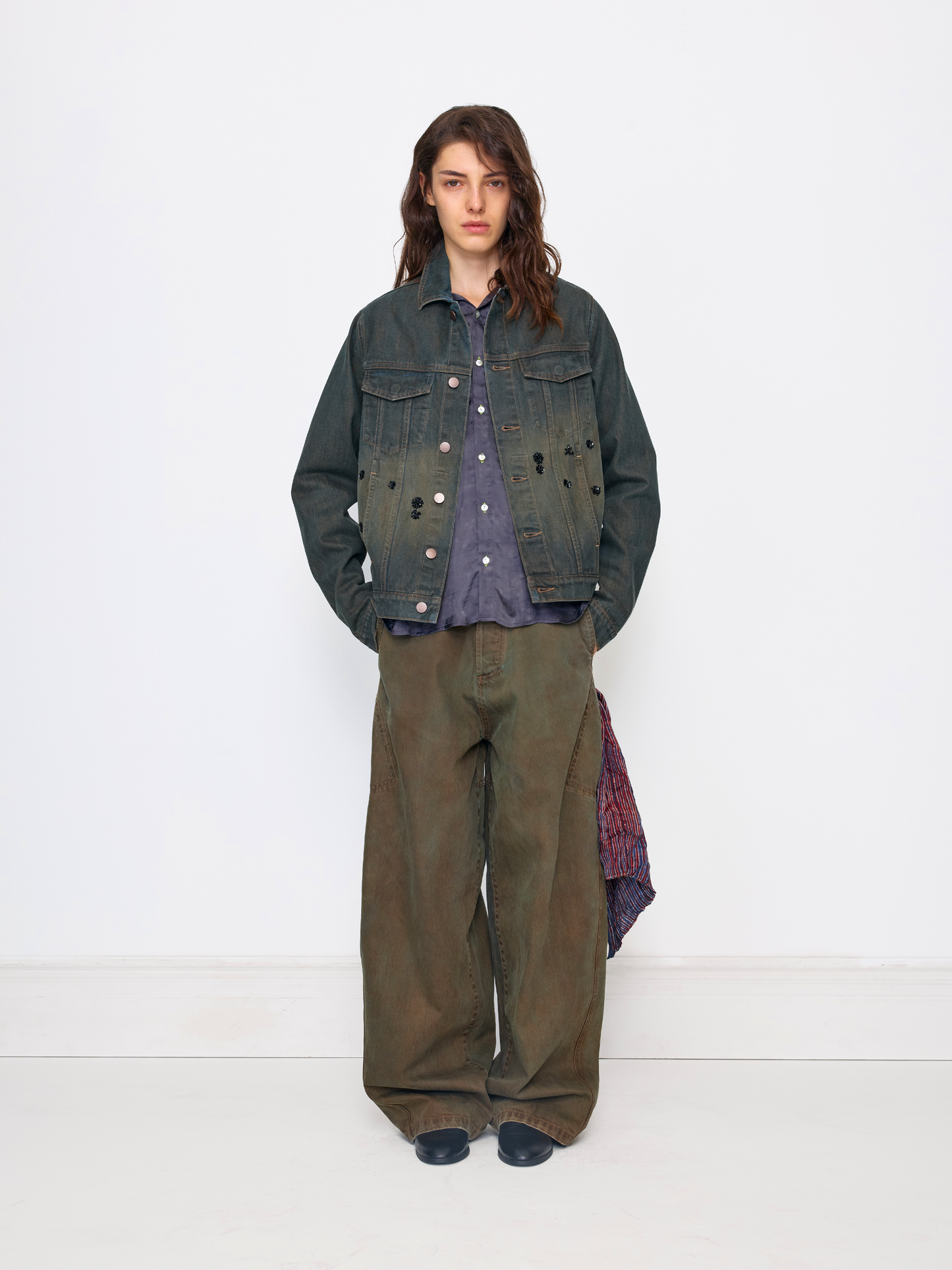 A model stands against a plain white studio wall wearing a dark distressed denim jacket over a purple button-up shirt, paired with wide-leg olive green pants and dark shoes.