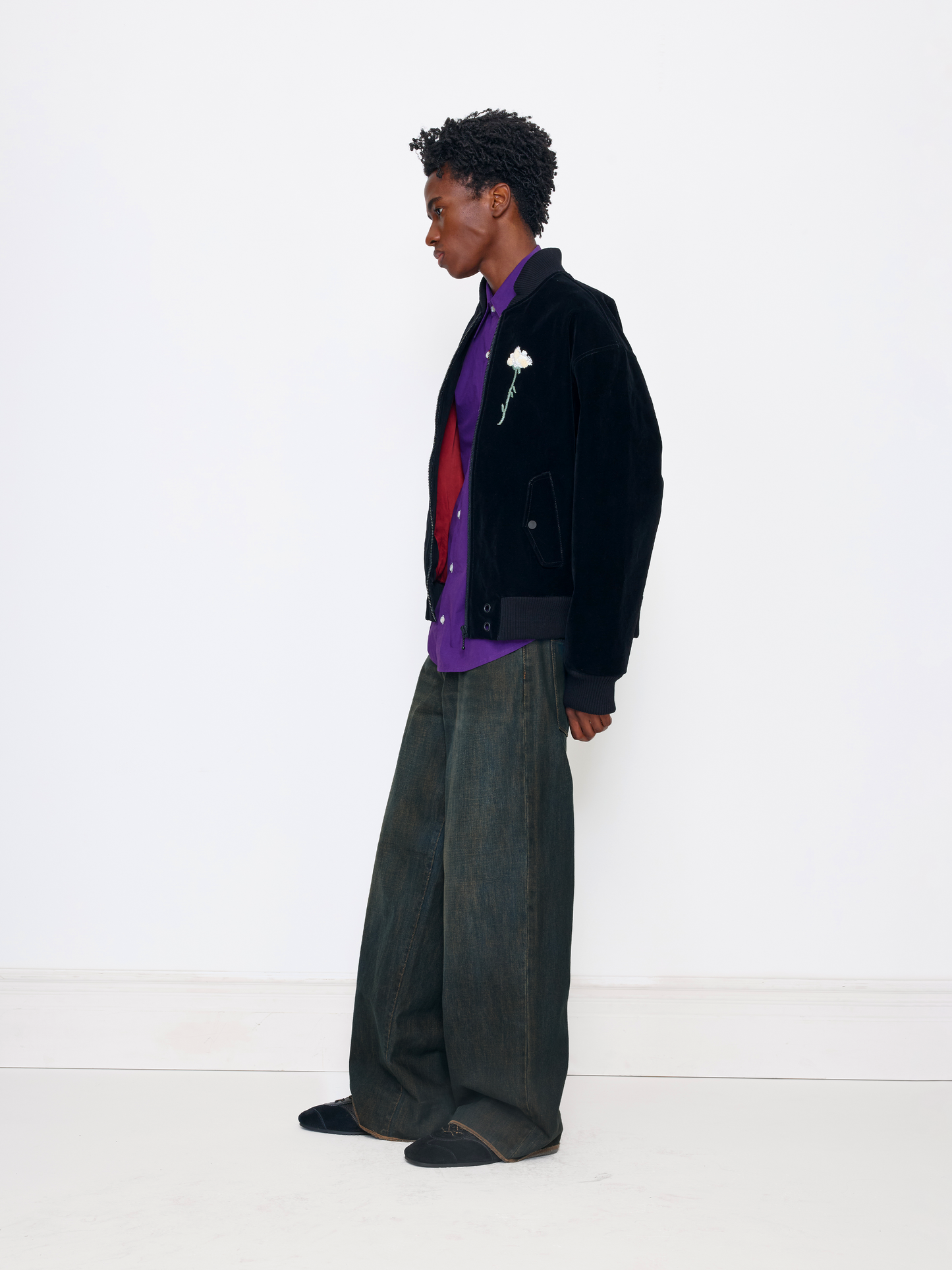 A model stands in profile wearing a black velvet jacket over a purple and red shirt, paired with wide-leg dark gray trousers and black shoes, against a clean white studio background.