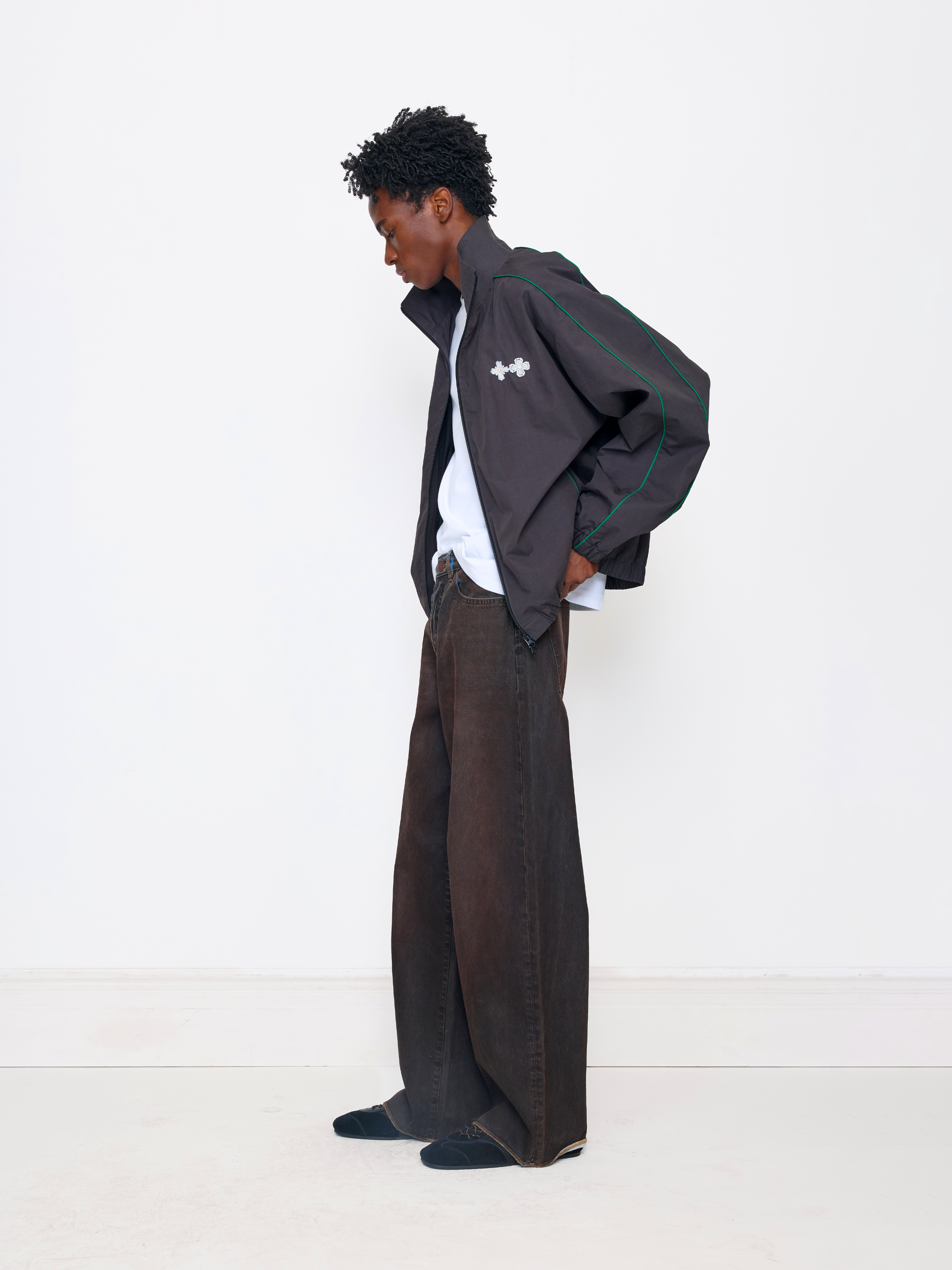 A model stands in profile wearing a dark gray zip-up jacket with green piping and a white embroidered logo, layered over a white shirt and paired with wide-leg brown trousers and dark loafers, against a clean white studio background.