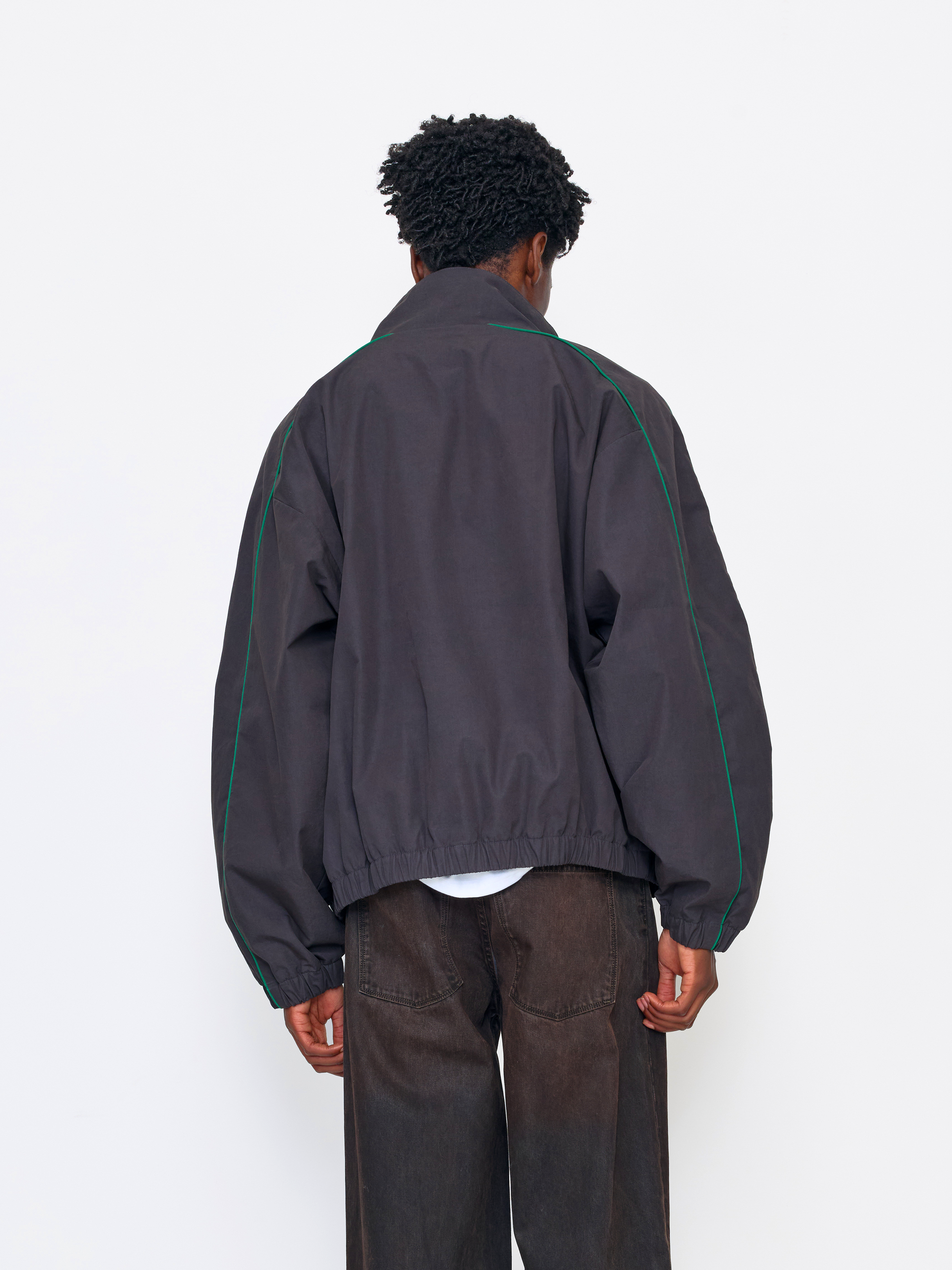 A person shown from the back wearing a dark gray windbreaker jacket with thin green piping along the shoulders and sleeves, paired with dark brown pants, against a plain white studio background.