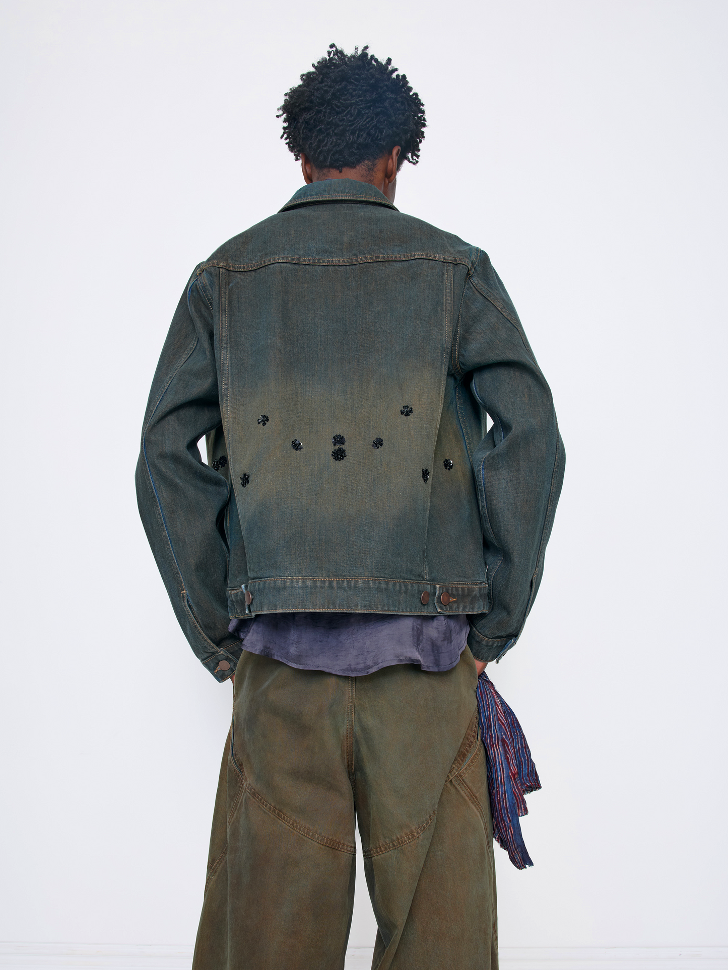 A model viewed from behind wearing a dark washed denim jacket with unique button details on the back, olive green wide-leg pants, and a purple shirt underneath, set against a plain white studio background.