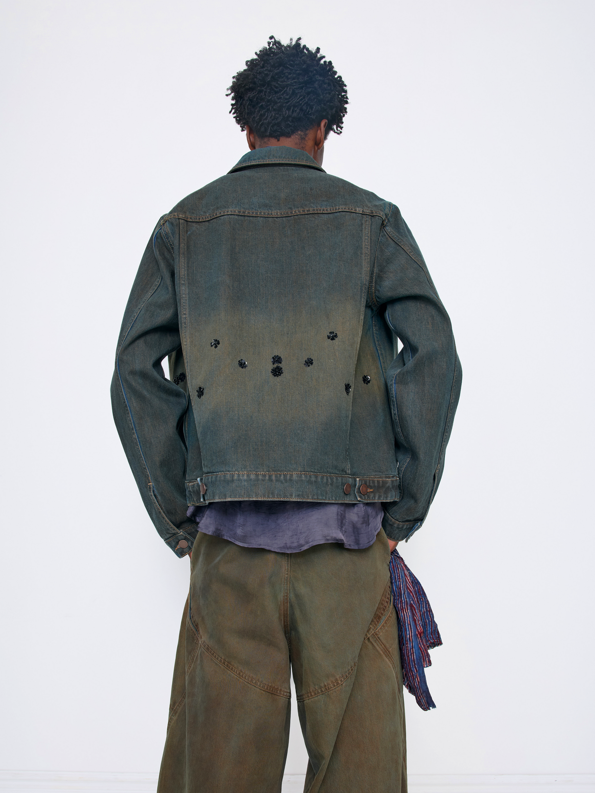 A model viewed from behind wearing a dark washed denim jacket with unique button details on the back, olive green wide-leg pants, and a purple shirt underneath, set against a plain white studio background.