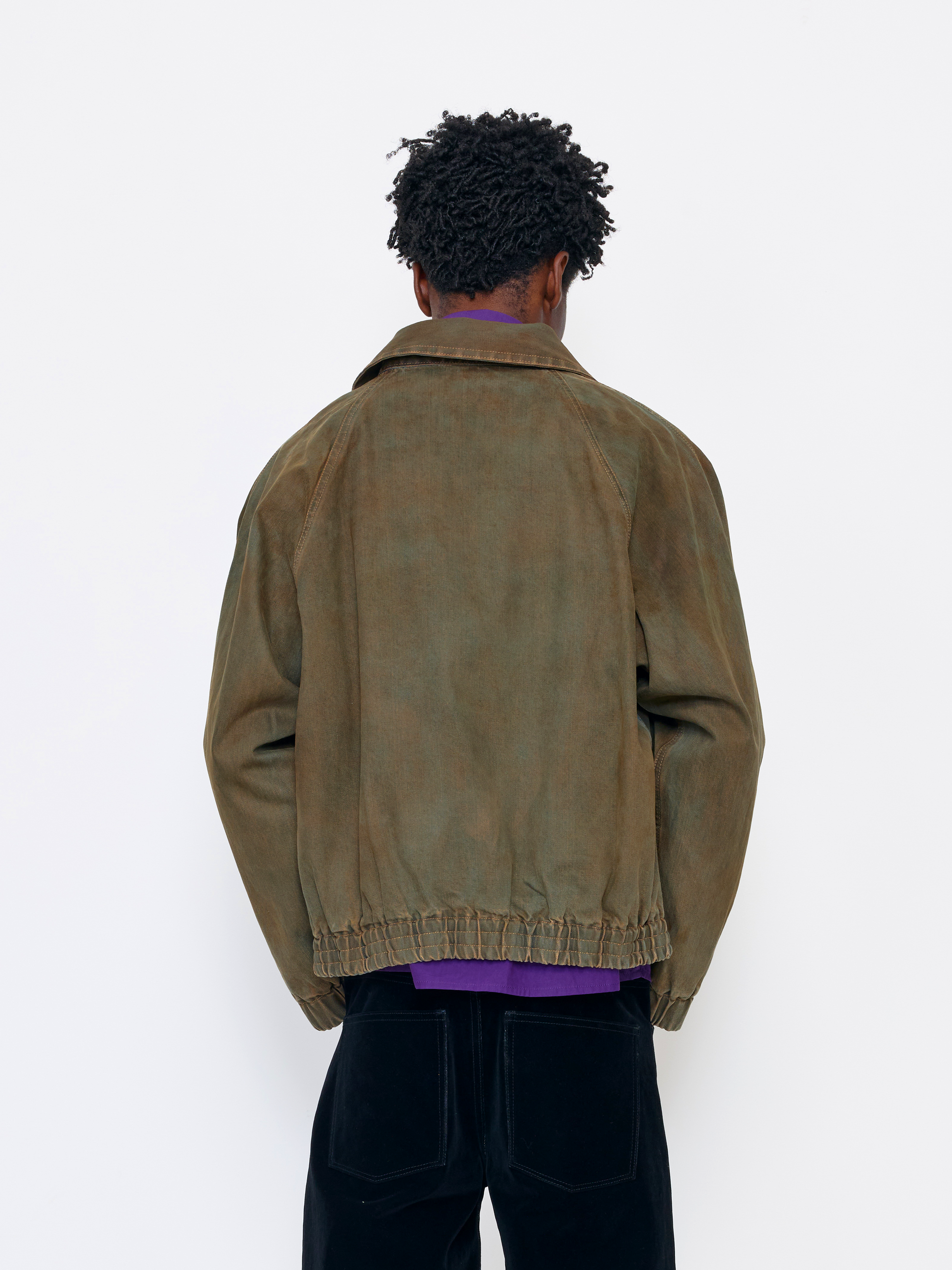 A person shown from the back wearing an oversized brown suede or washed leather jacket over a purple shirt and black pants, standing against a plain white wall.