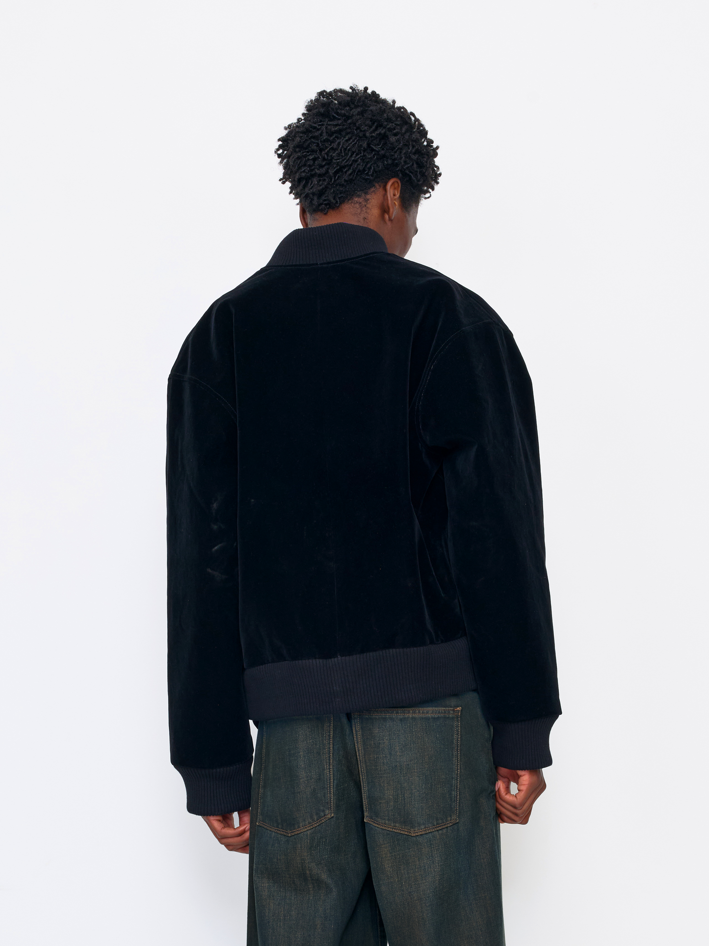 A person shown from the back wearing a black velvet jacket with ribbed collar, cuffs, and hem, paired with dark blue jeans, against a plain white studio background.