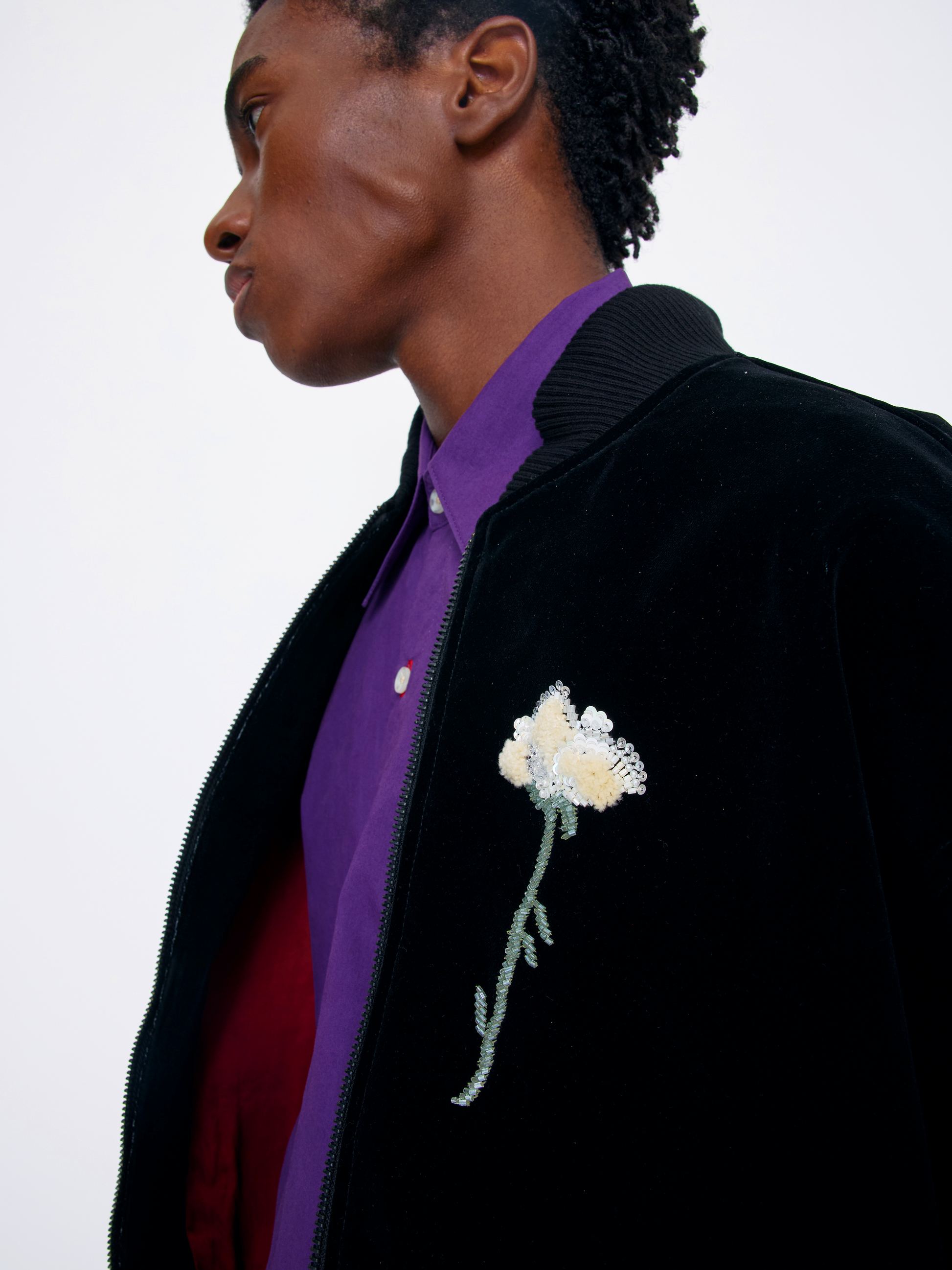 A close-up side profile of a model wearing a black velvet bomber jacket with a white and green sequined rose brooch on the chest, layered over a purple shirt and a red sweater, against a plain white background.