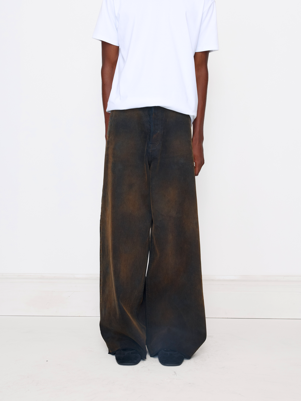 A person shown from the back wearing very wide-leg dark brown pants with a faded, mottled texture, paired with a plain white t-shirt, against a clean white studio background.