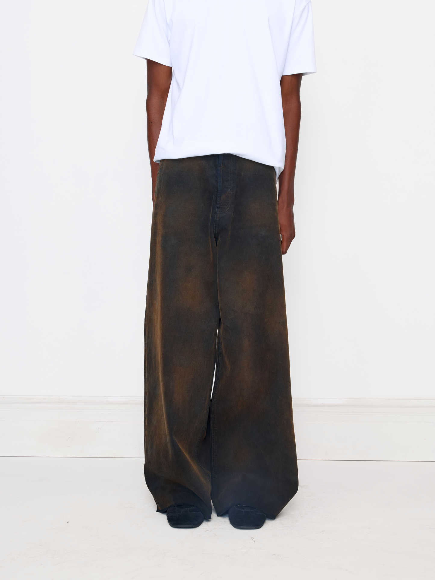 A person shown from the back wearing very wide-leg dark brown pants with a faded, mottled texture, paired with a plain white t-shirt, against a clean white studio background.