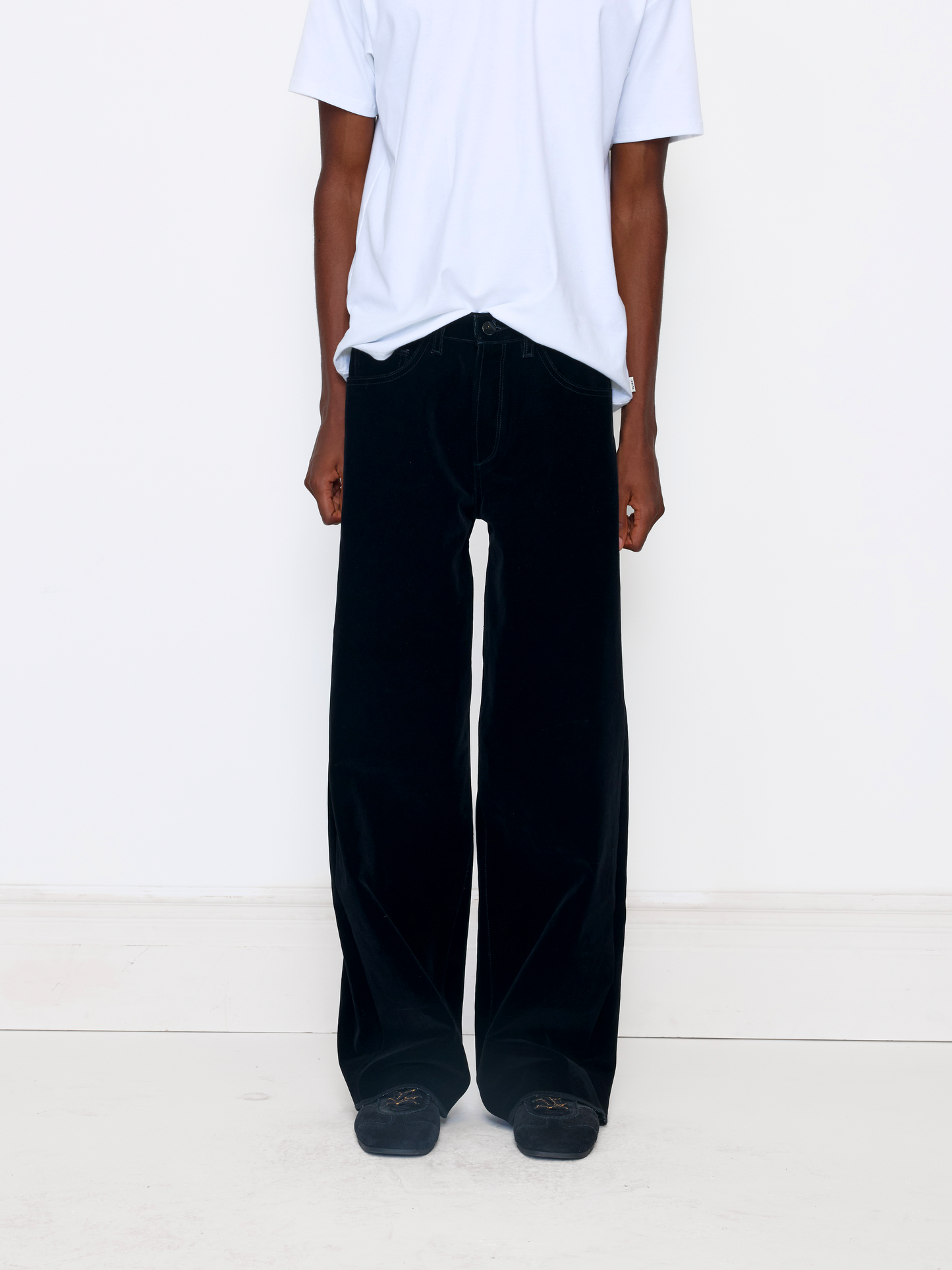 A model wearing black wide-leg velvet trousers, paired with a white t-shirt and black shoes, standing against a plain white studio background.