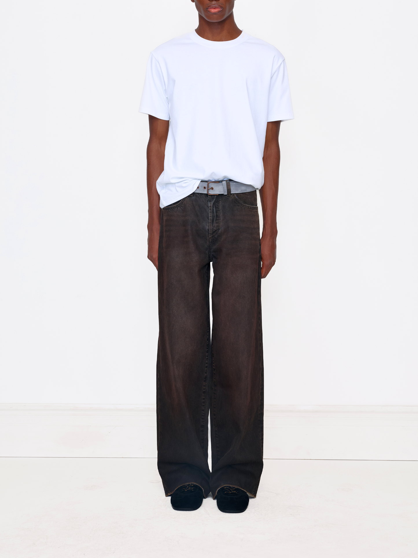 A model wearing a white crewneck t-shirt tucked into dark brown wide-leg jeans with a gray belt, standing against a plain white studio background.