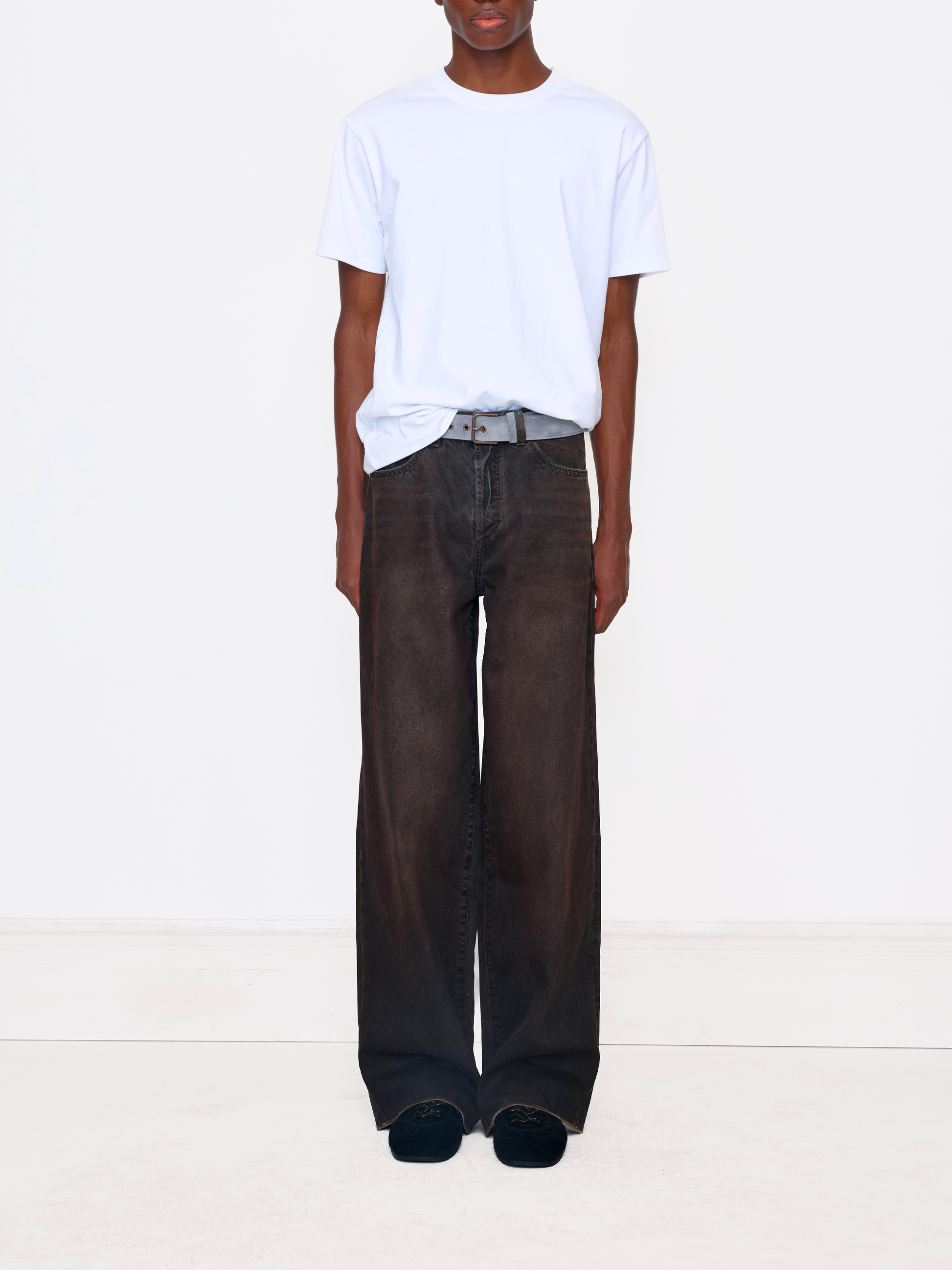 A model wearing a white crewneck t-shirt tucked into dark brown wide-leg jeans with a gray belt, standing against a plain white studio background.