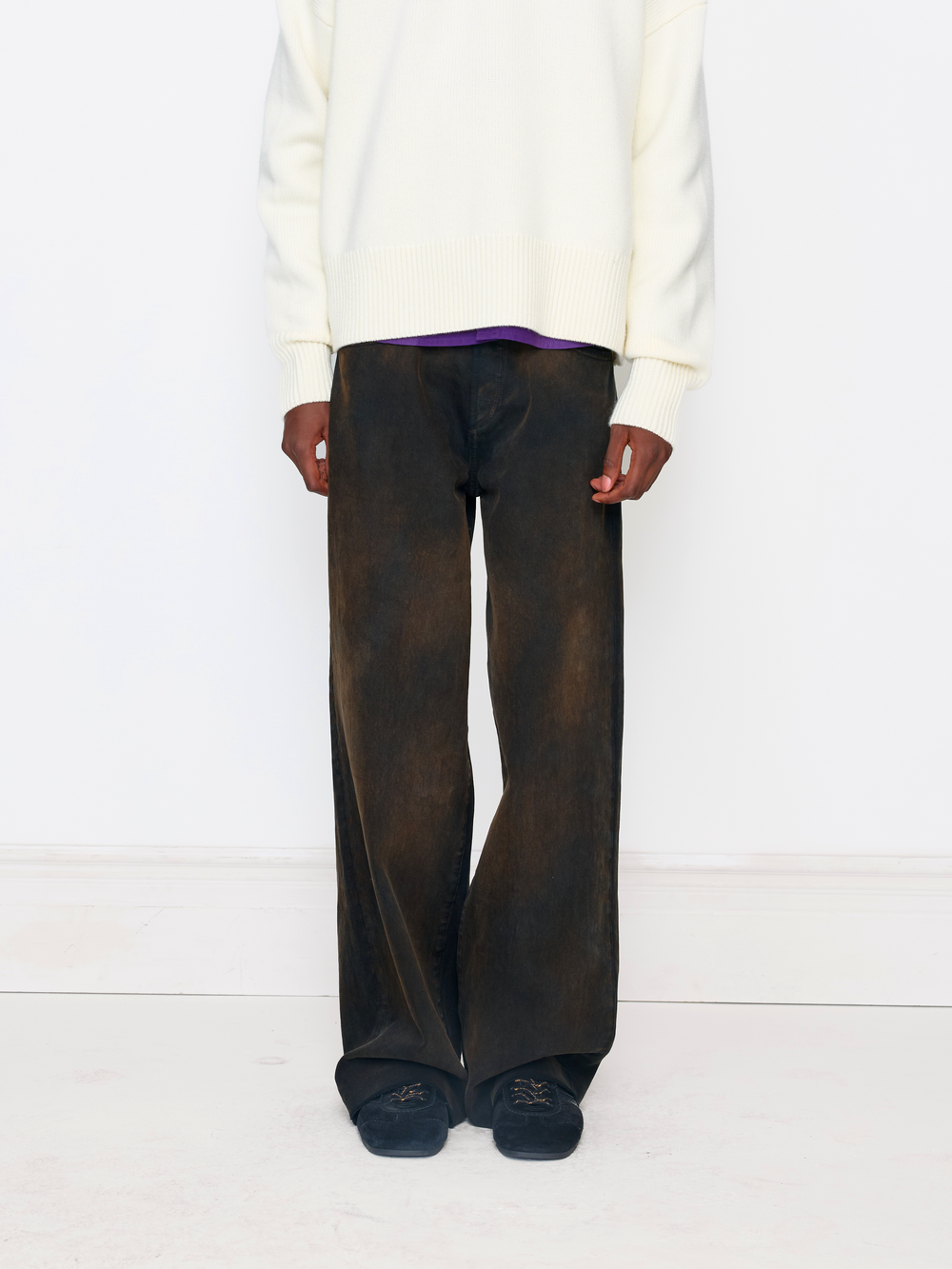 A model is shown from the chest down wearing wide-leg dark brown washed trousers, a cream knit sweater, a purple shirt, and dark slip-on shoes against a clean white studio background.