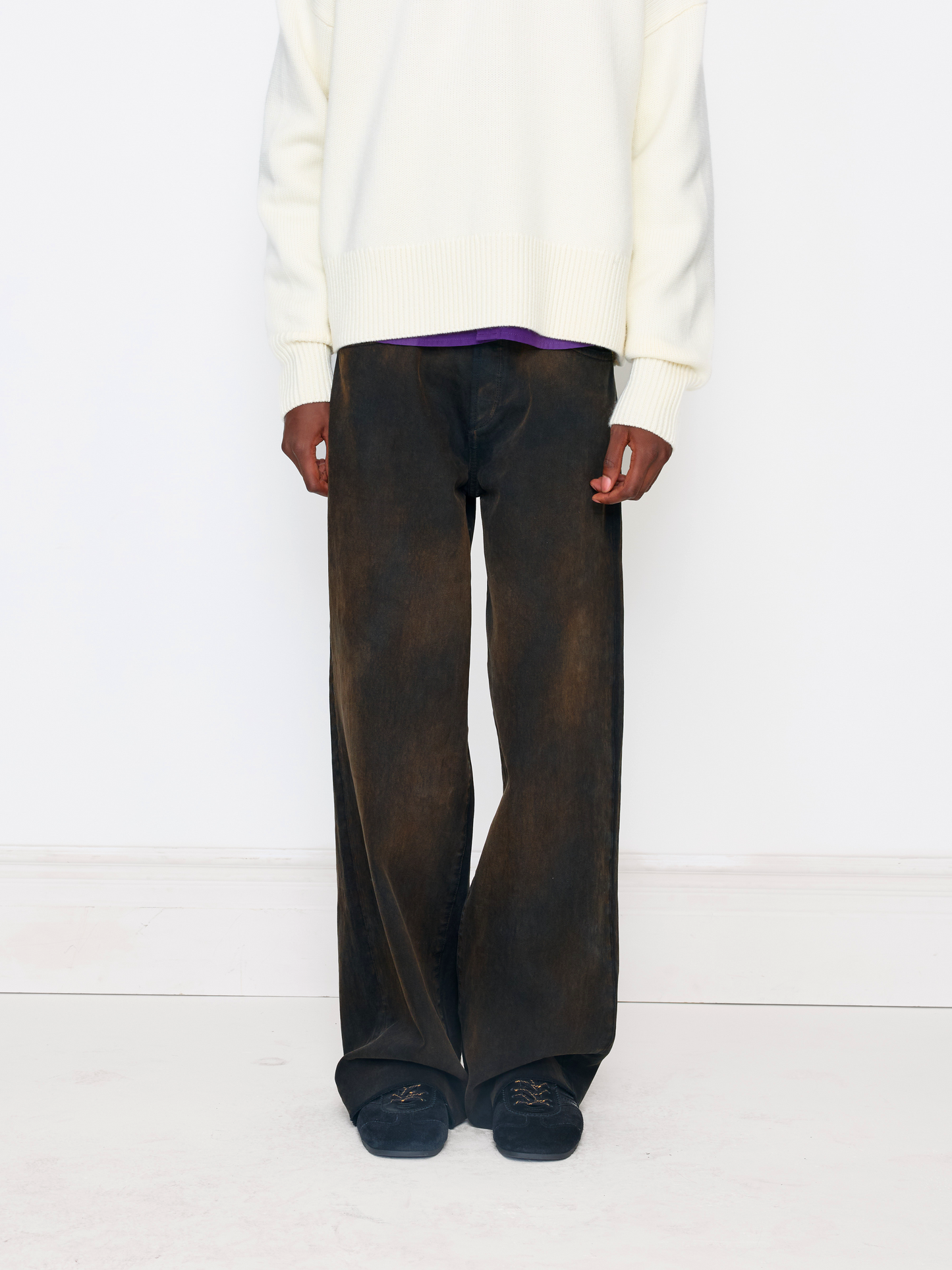 A model is shown from the chest down wearing wide-leg dark brown washed trousers, a cream knit sweater, a purple shirt, and dark slip-on shoes against a clean white studio background.