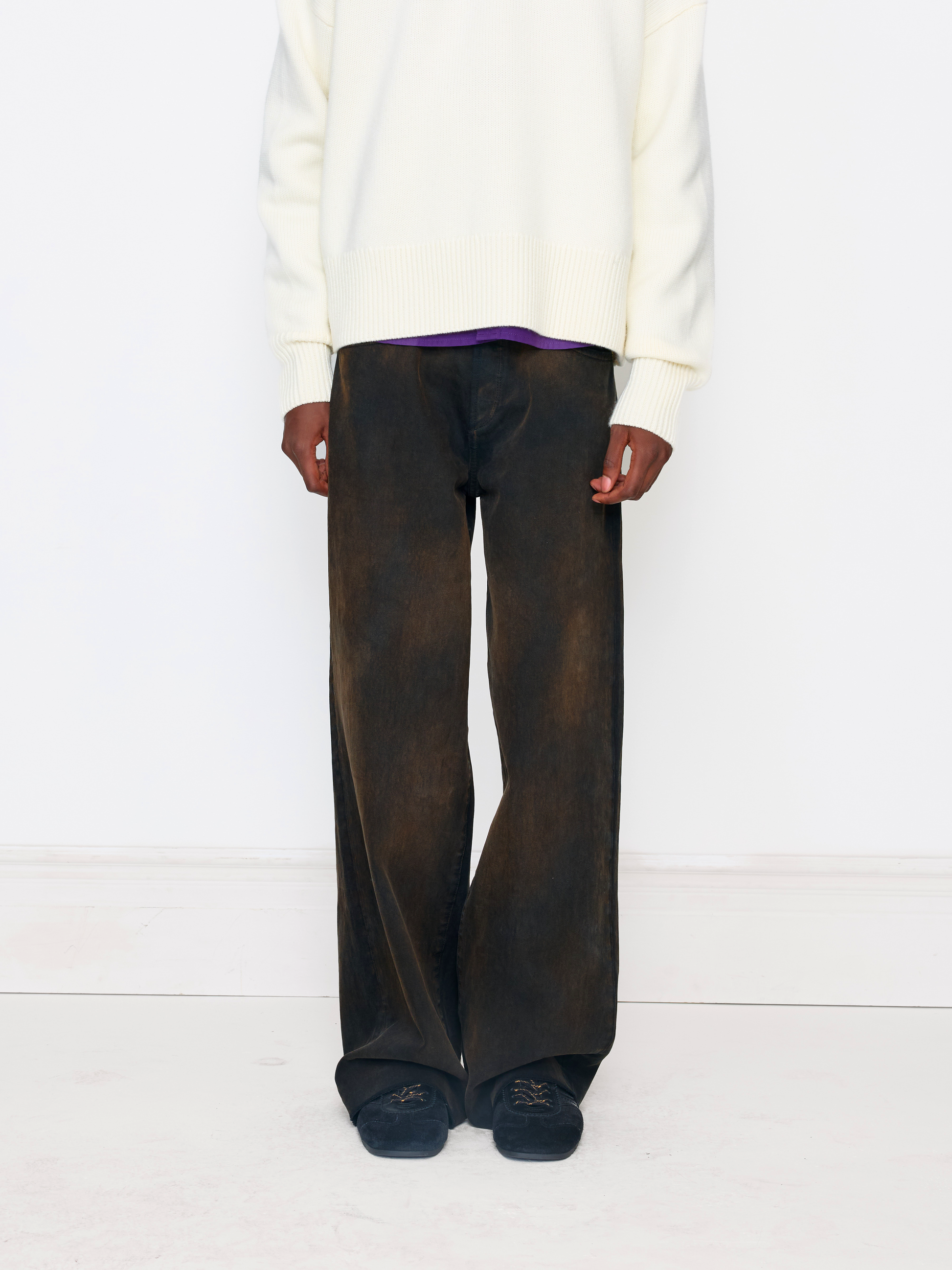 A model is shown from the chest down wearing wide-leg dark brown washed trousers, a cream knit sweater, a purple shirt, and dark slip-on shoes against a clean white studio background.