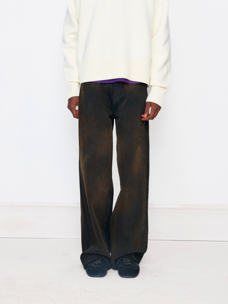 A model is shown from the chest down wearing wide-leg dark brown washed trousers, a cream knit sweater, a purple shirt, and dark slip-on shoes against a clean white studio background.