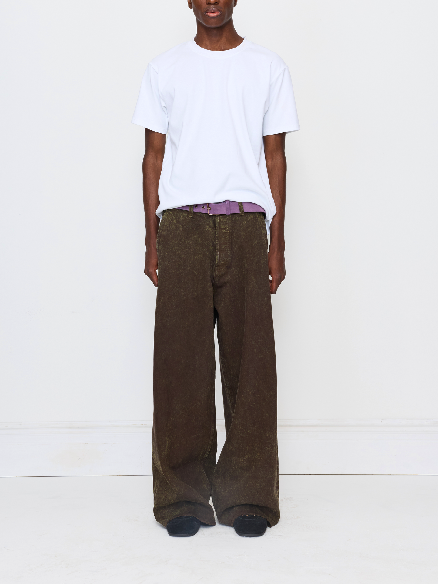 A model wearing a white crewneck t-shirt, brown wide-leg corduroy trousers, a purple patterned belt, and black slip-on shoes, standing against a plain white wall.