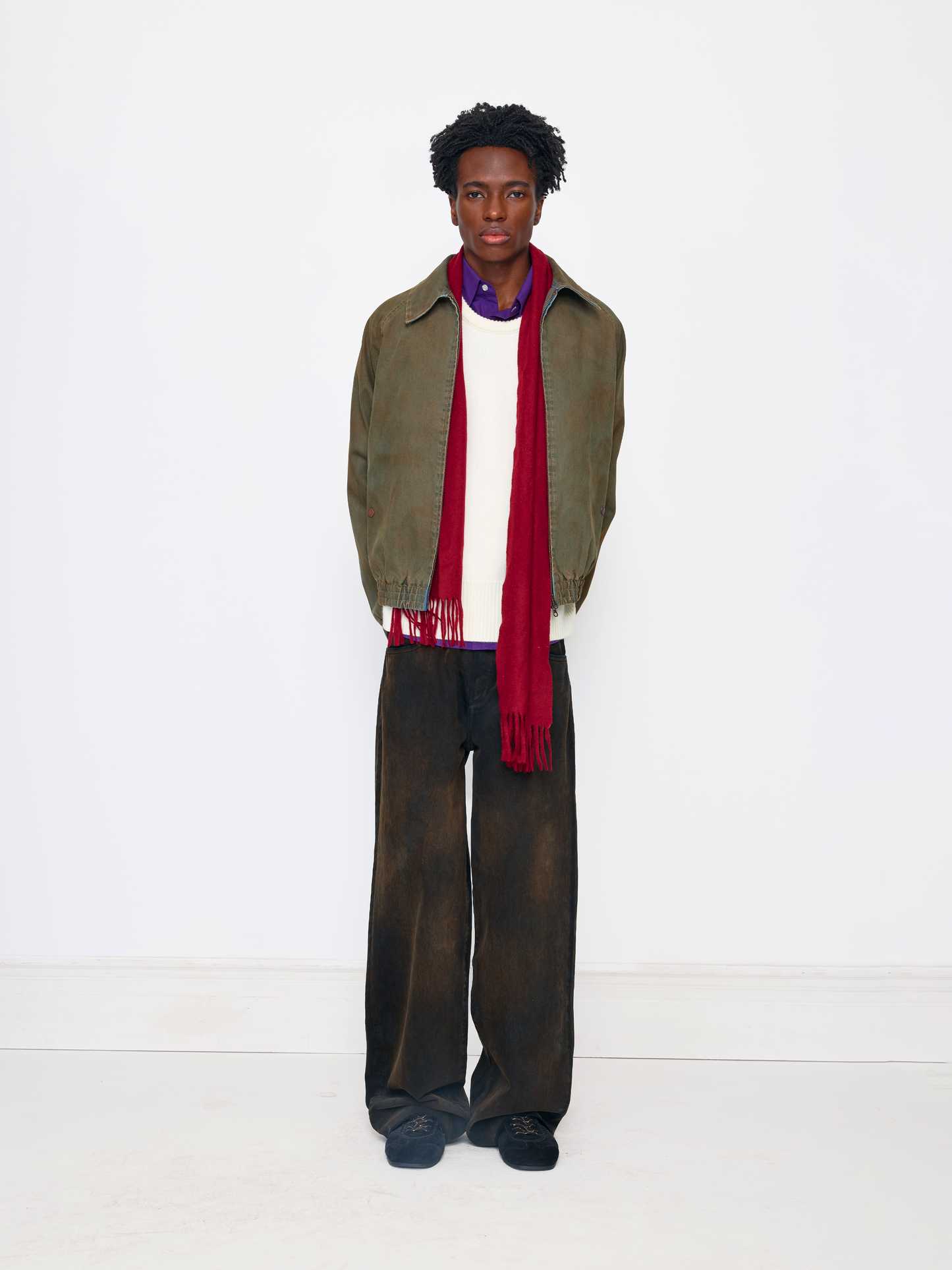 A model stands against a plain white studio background wearing an olive green jacket, a white sweater, a red scarf, dark brown wide-leg trousers, and dark blue shoes.