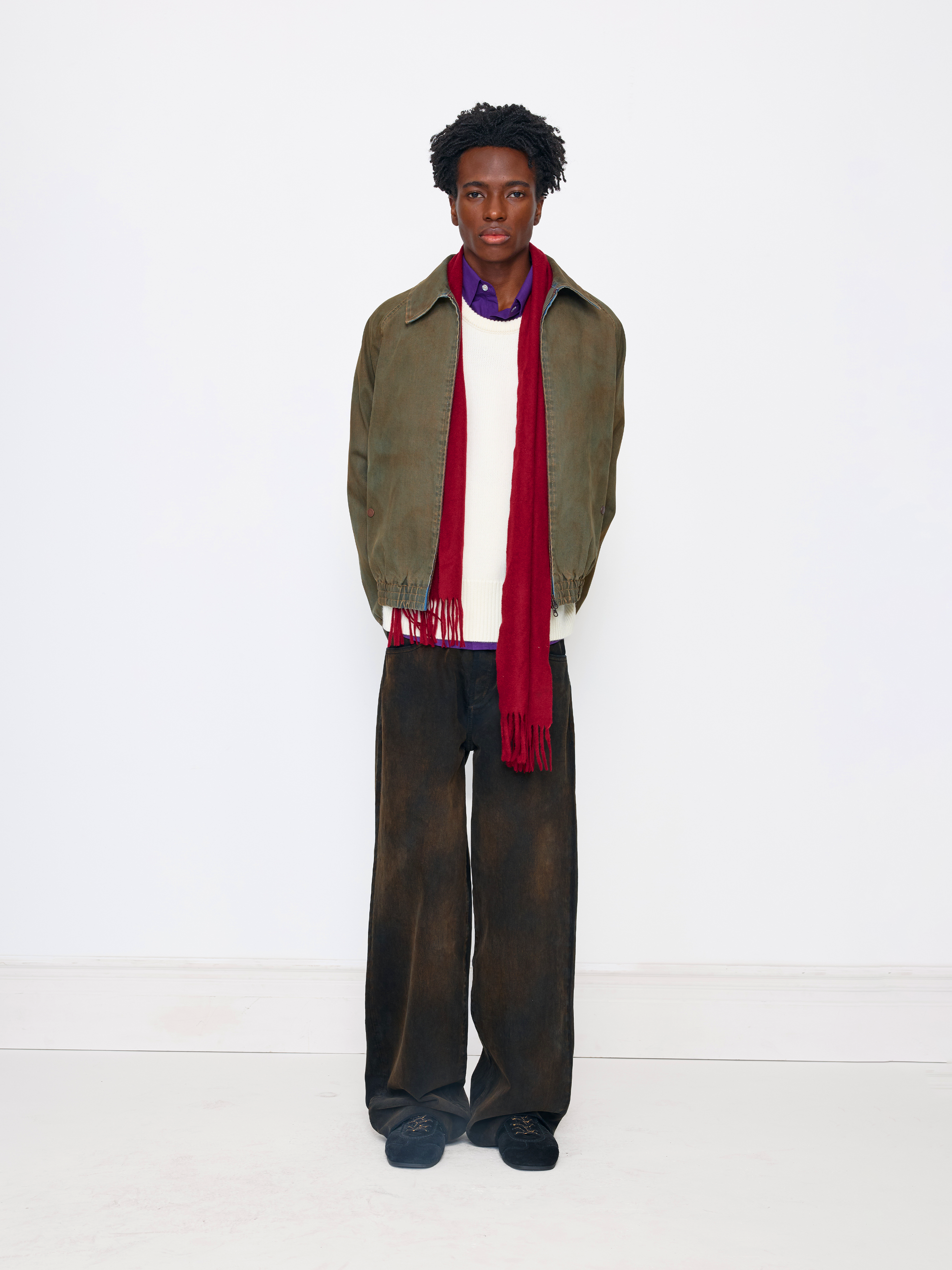 A model stands against a plain white studio background wearing an olive green jacket, a white sweater, a red scarf, dark brown wide-leg trousers, and dark blue shoes.