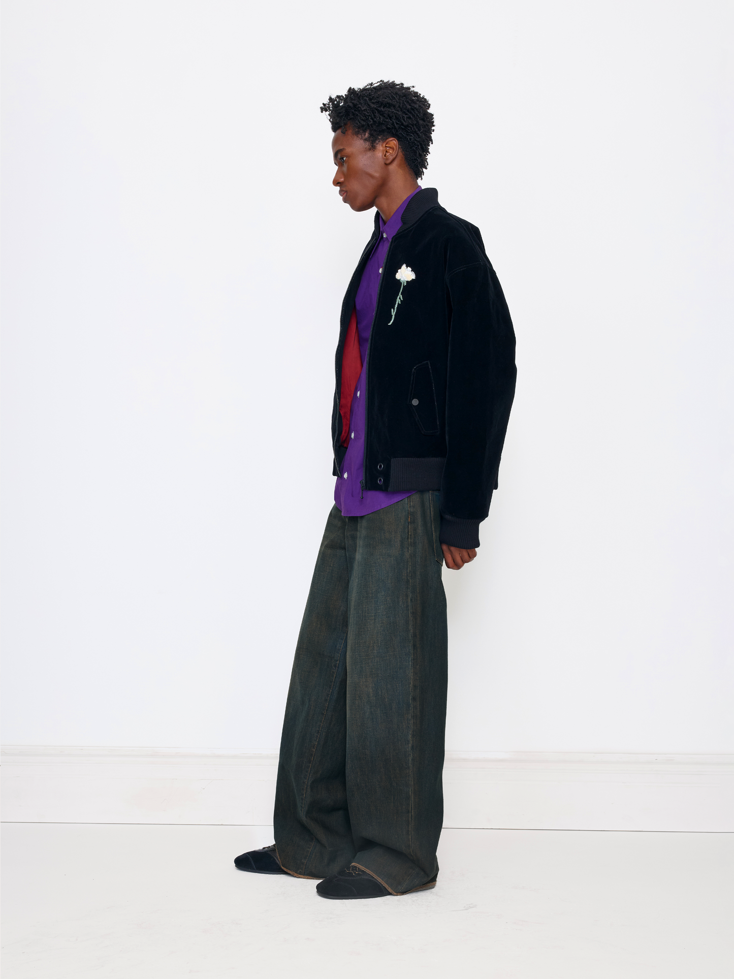 A model stands in profile wearing a black velvet jacket with a white rose lapel pin, layered over a purple shirt and red sweater, paired with wide-leg dark gray jeans and black slip-on shoes, against a clean white studio background.