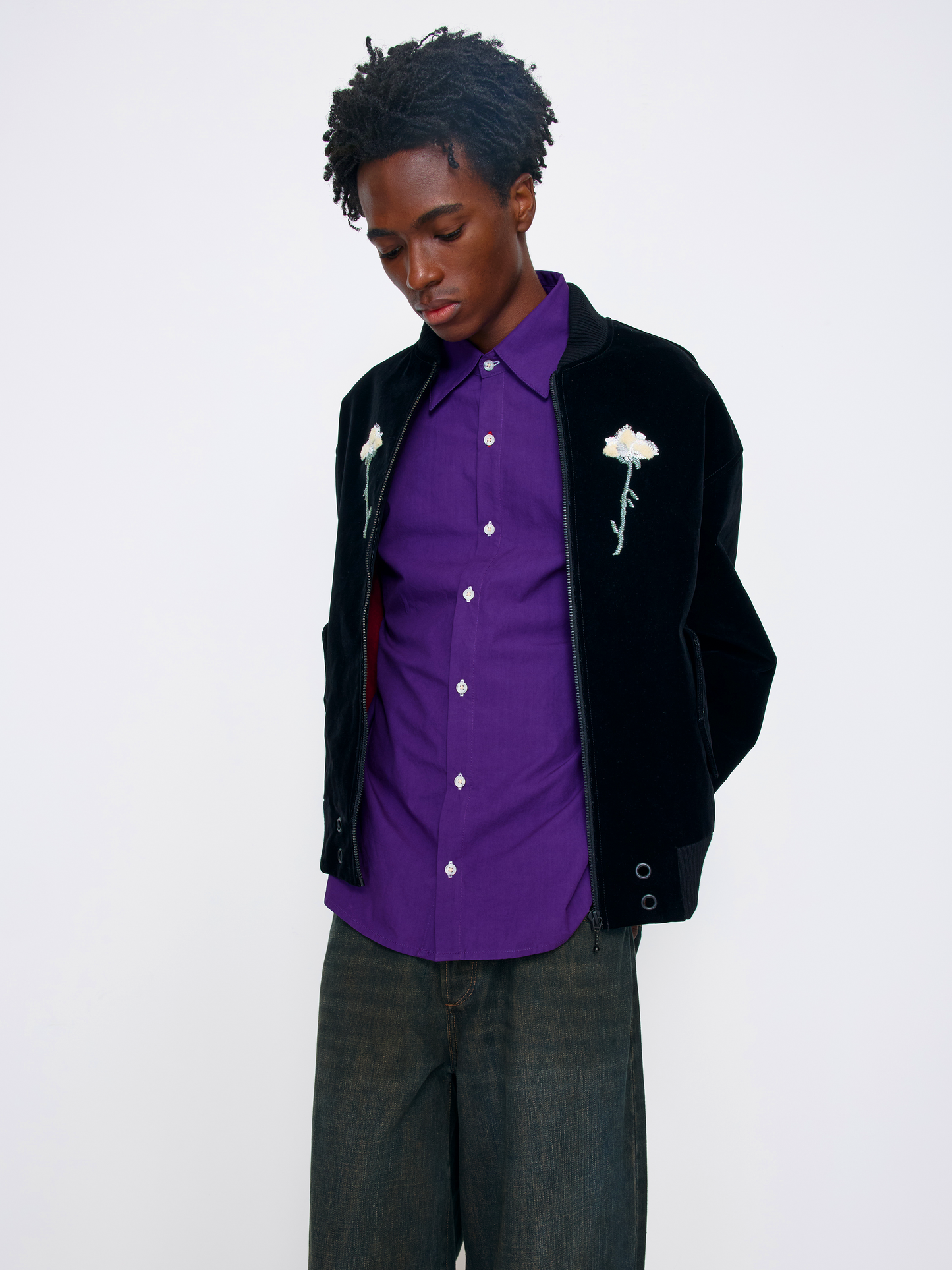 A model wearing a black velvet bomber jacket with white embroidered roses, a purple button-down shirt, and dark gray jeans, photographed against a plain white studio background.