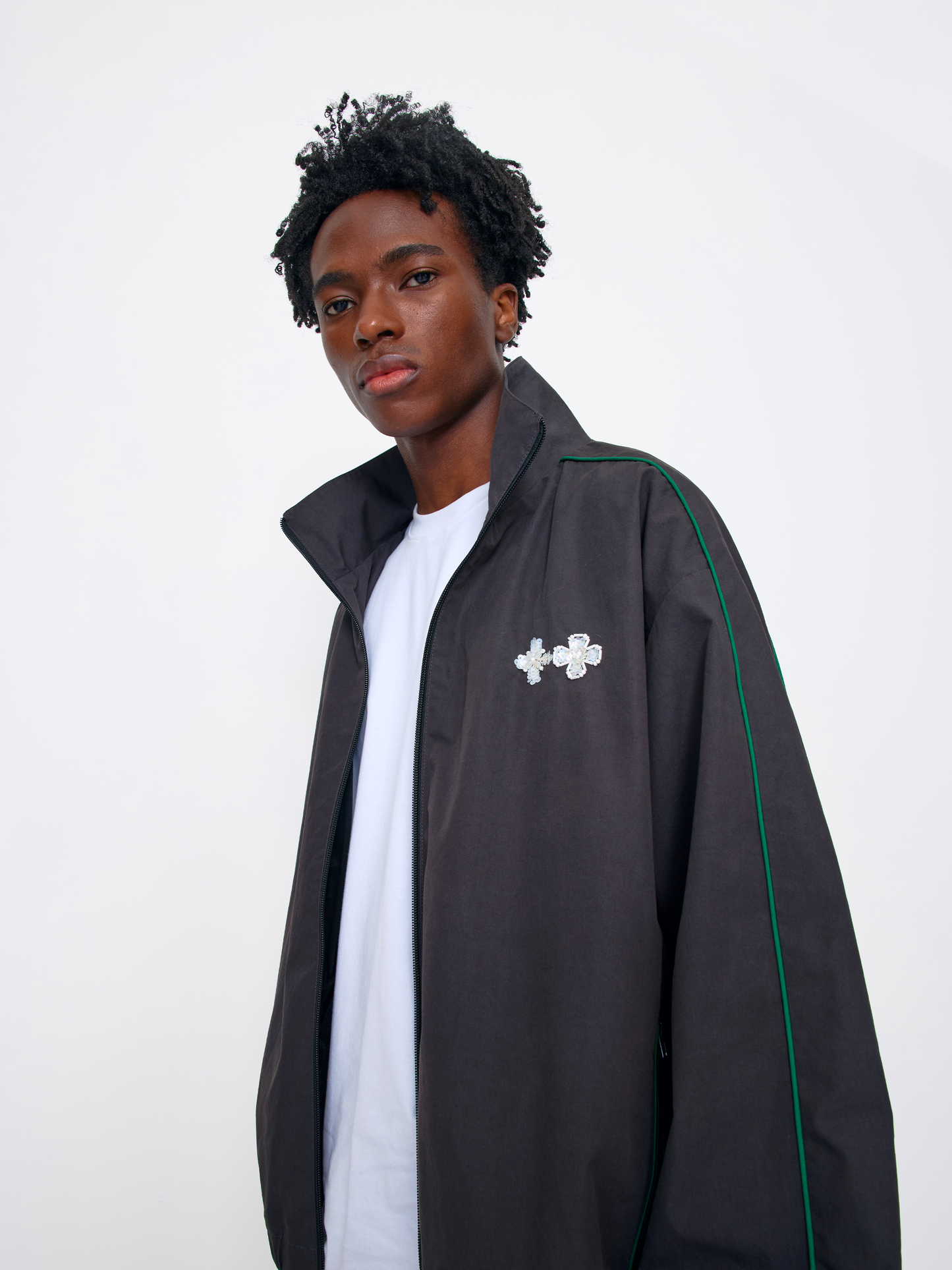 A young man with curly black hair wearing a dark gray zip-up track jacket with green piping and a silver cross brooch, layered over a white t-shirt, against a plain white studio background.