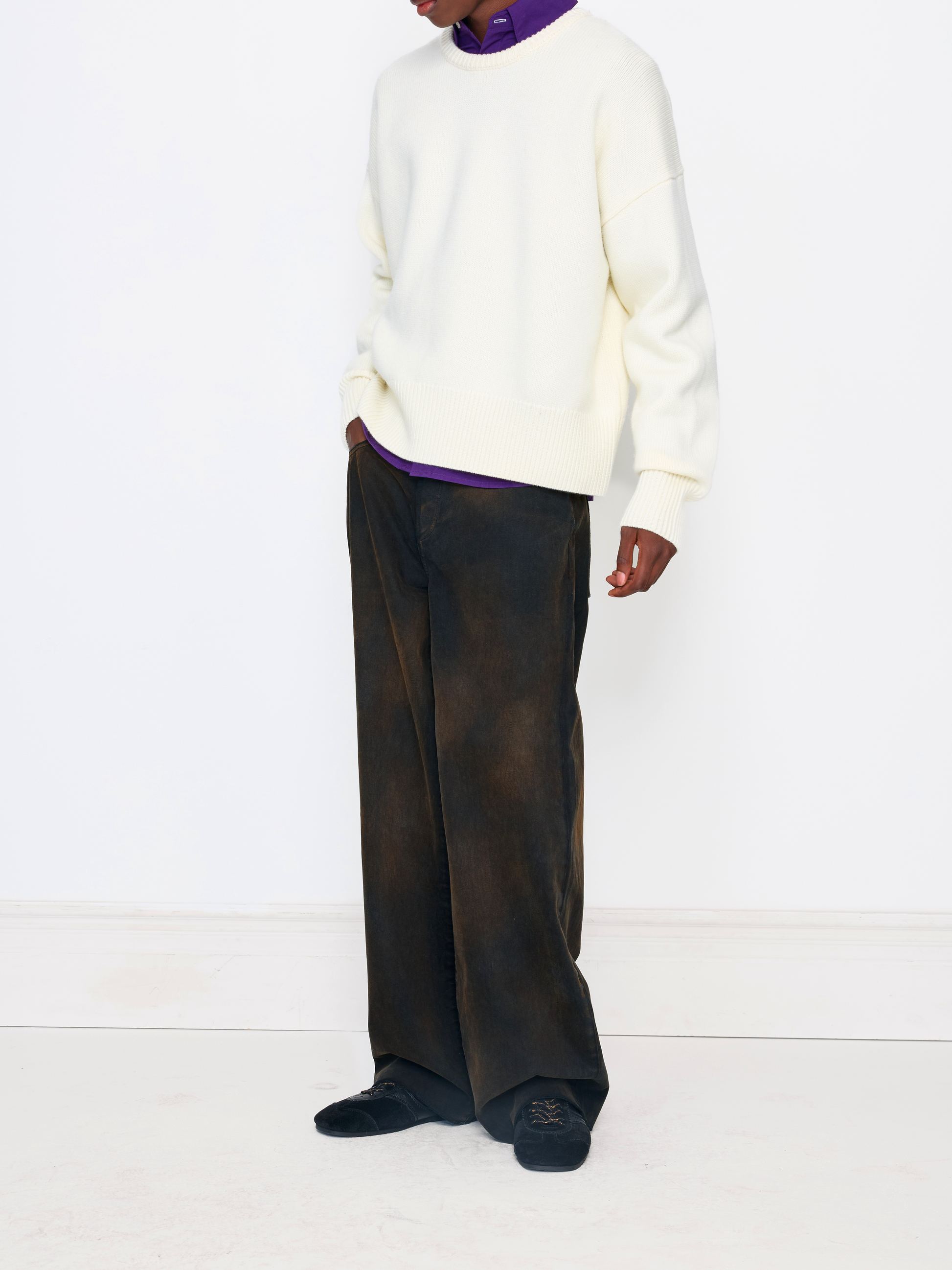 A model wearing a cream-colored crewneck sweater, dark brown wide-leg trousers, and black shoes, styled with a purple turtleneck underneath the sweater, set against a plain white studio background.