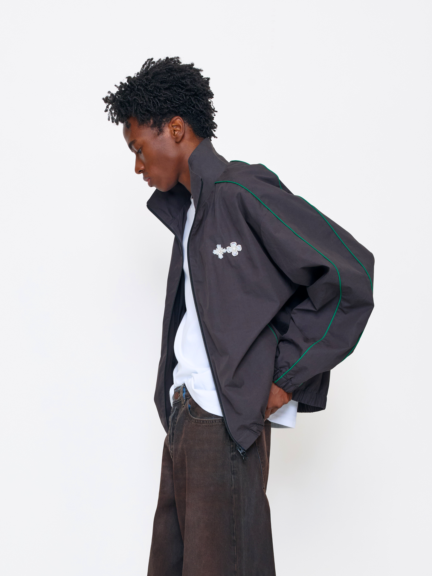 A model wearing a dark gray zip-up jacket with green piping and a white cross logo on the chest, styled over a white t-shirt and brown pants, set against a plain white studio background.