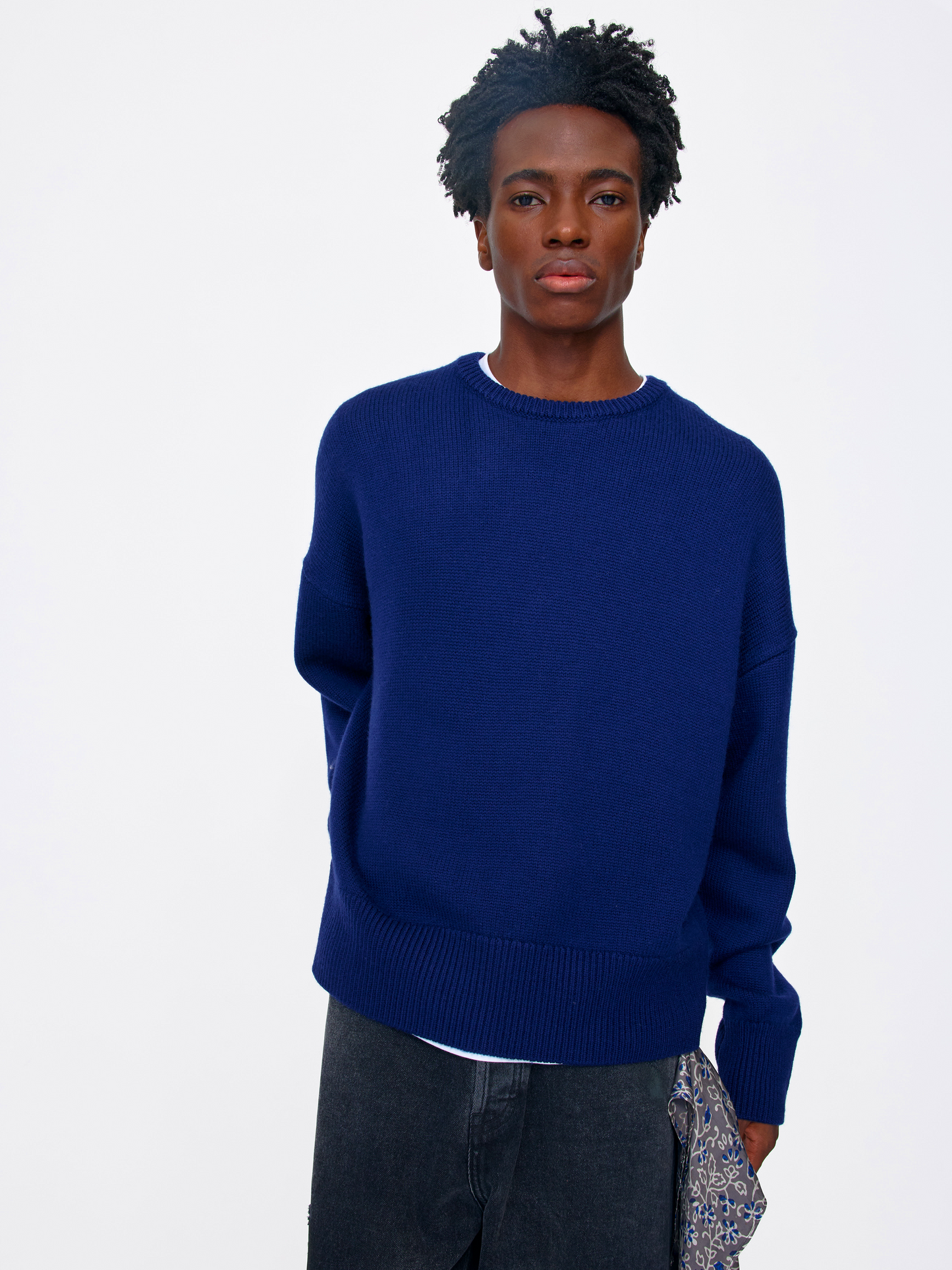 A man wearing a solid navy blue crewneck knit sweater, dark gray jeans, and holding a patterned clutch bag, photographed against a clean white studio background.