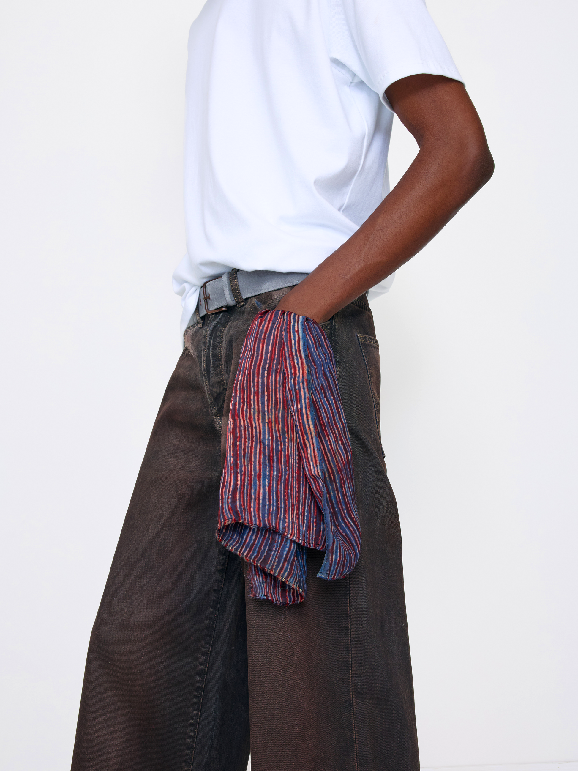 A model wearing a white t-shirt, dark brown wide-leg jeans, and a white belt, with a red, blue, and purple striped button-down shirt tucked into the back pocket.