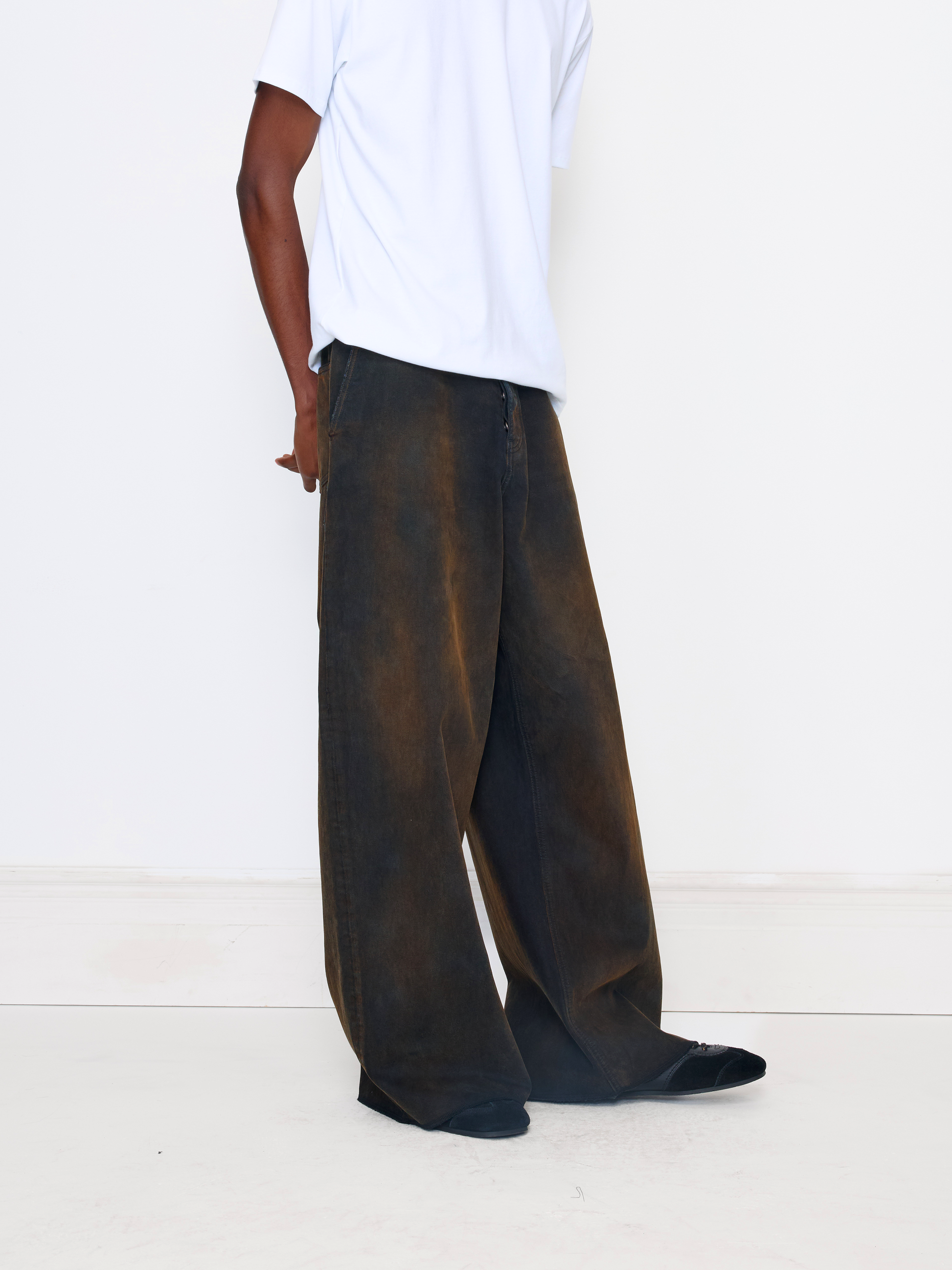 A person wearing very wide-leg dark brown pants with a faded, washed texture, paired with a white t-shirt and black shoes, standing against a plain white wall.