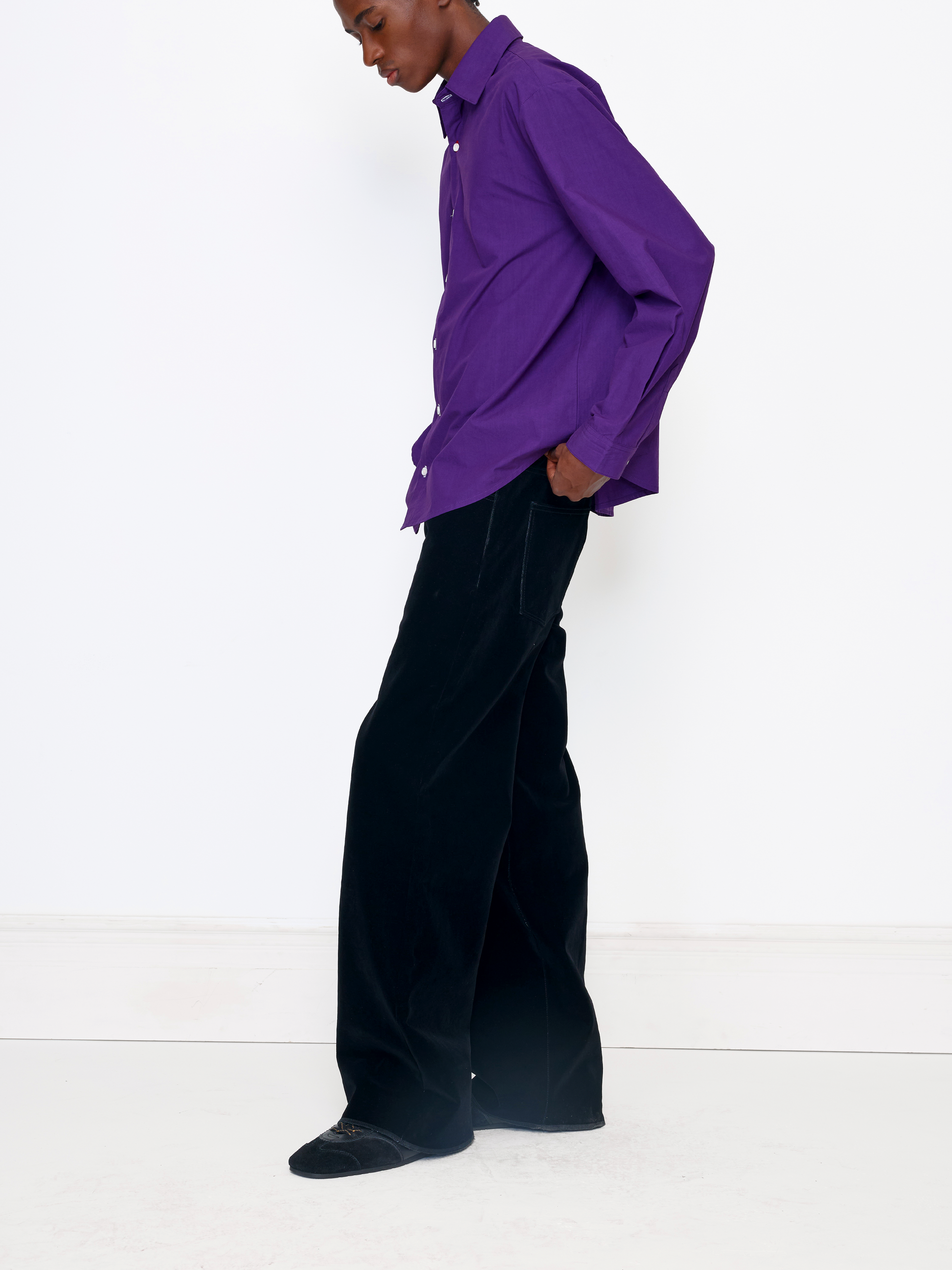 A model wearing a vibrant purple button-down shirt and black wide-leg trousers, standing in profile against a clean white studio background.