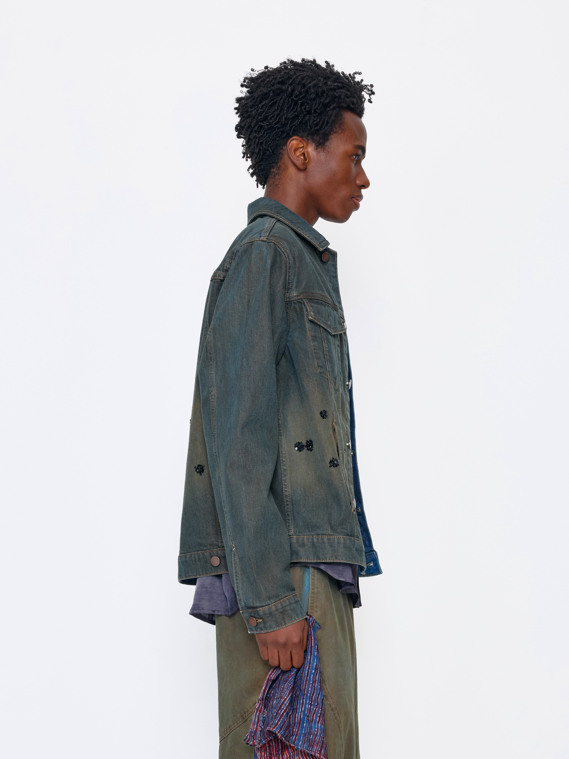A model shown in profile wearing a dark, washed denim jacket over a blue shirt, paired with olive pants and holding a striped fabric accessory, set against a plain white studio background.