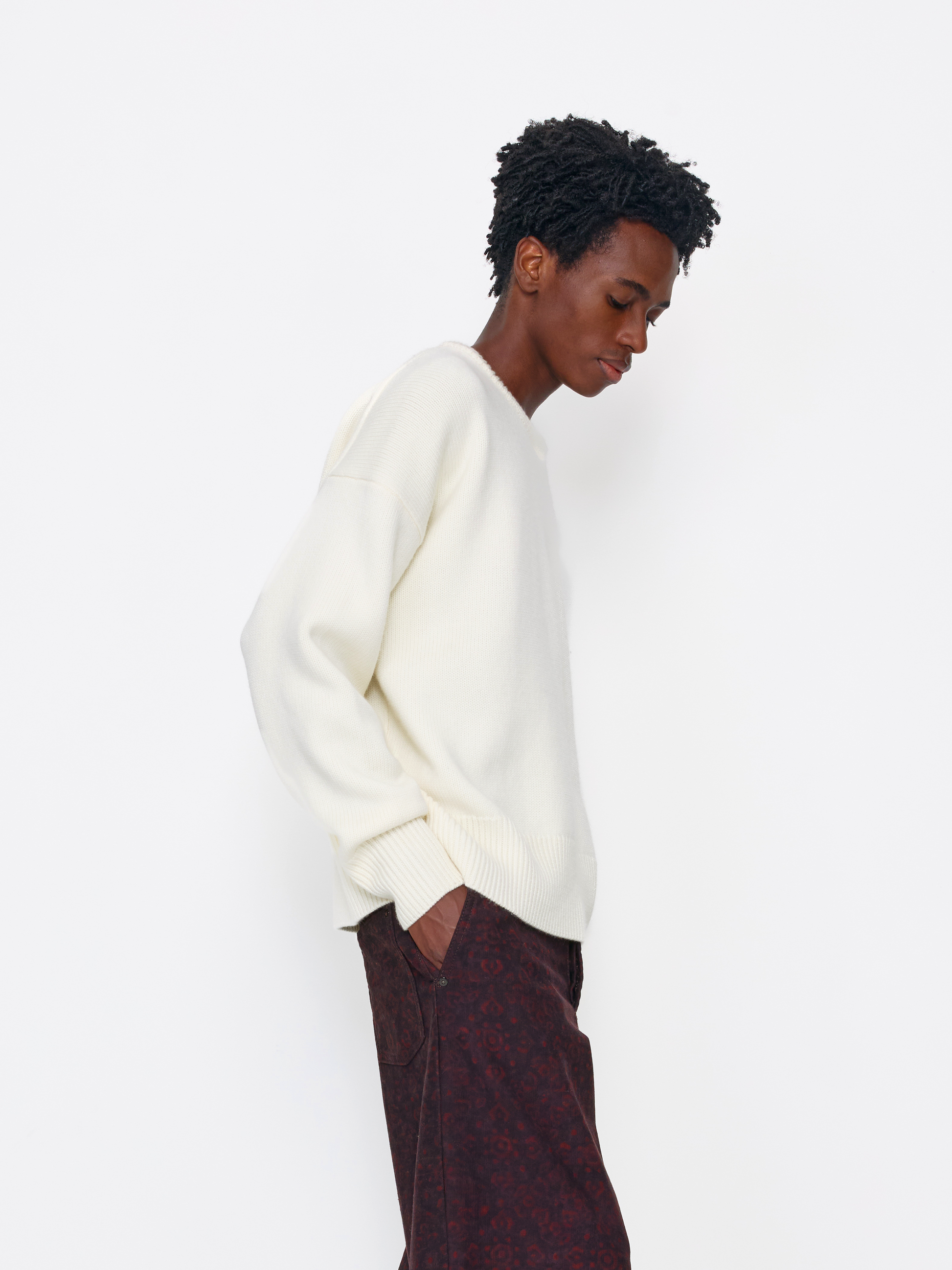 A model wearing an off-white ribbed knit crewneck sweater and dark patterned trousers, shown in profile against a plain white studio background.