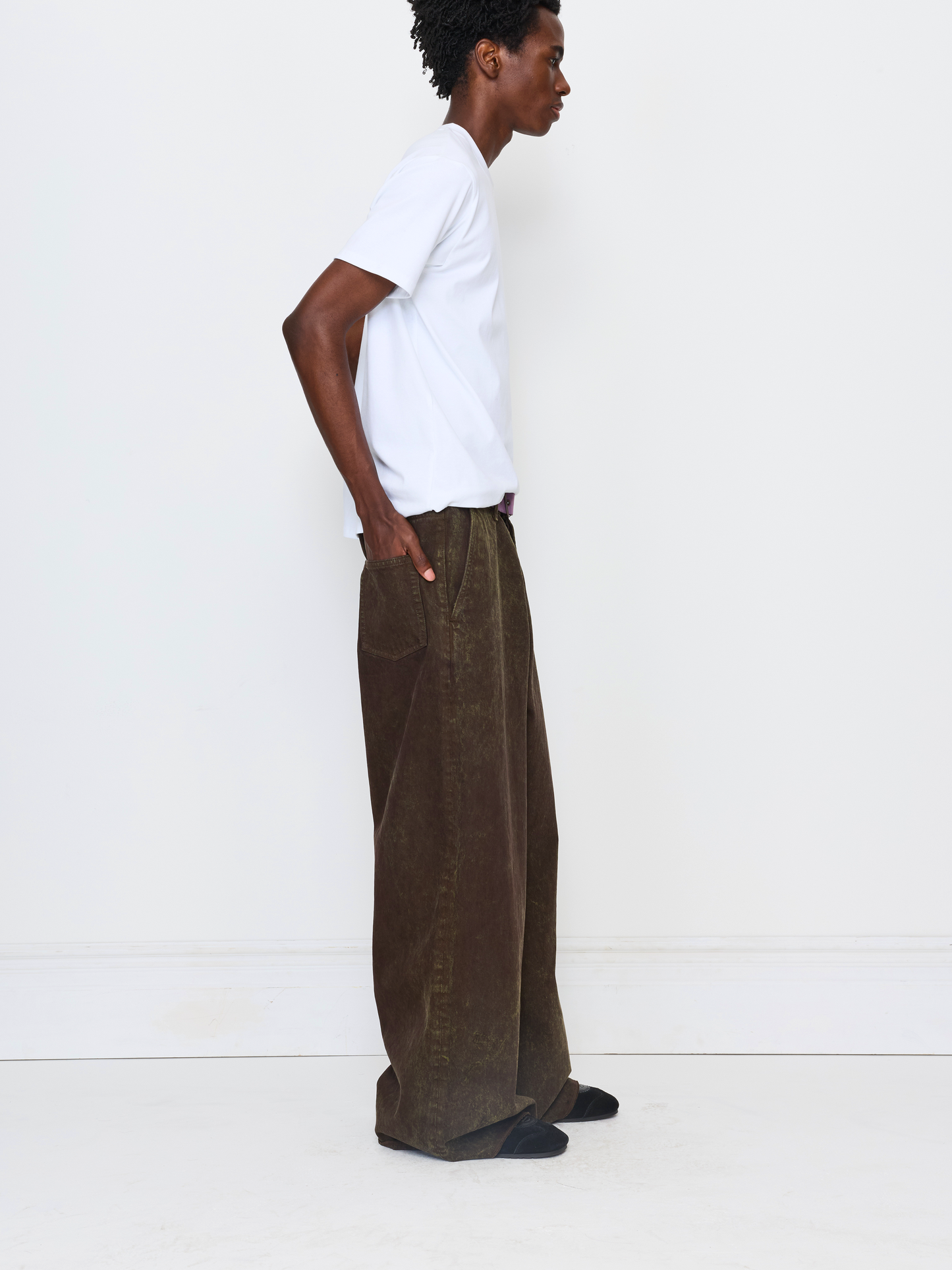 A model shown in profile wearing wide-leg brown corduroy trousers, a white t-shirt, and black shoes against a clean white studio background.