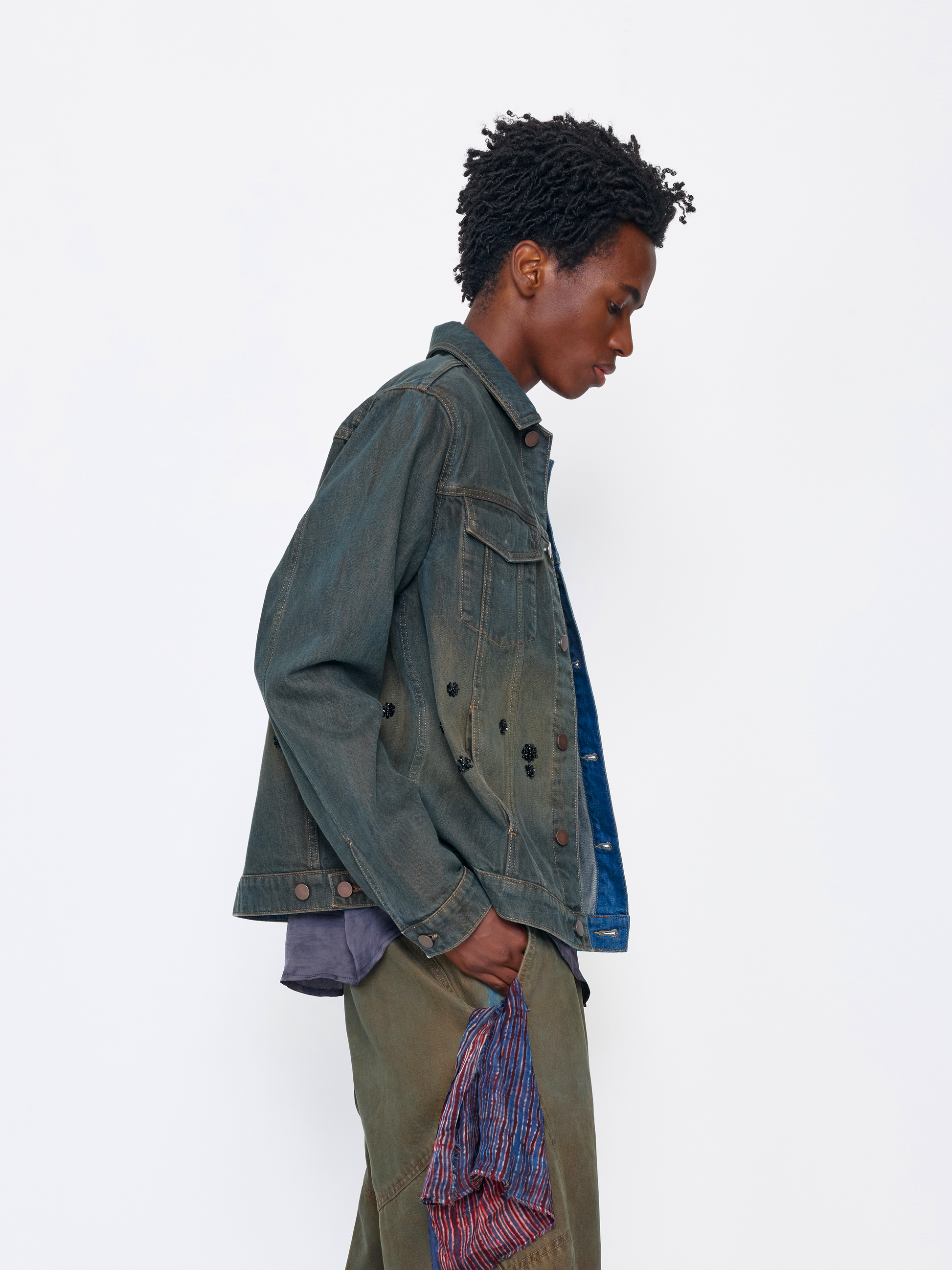 A model shown in profile wearing a dark washed denim jacket over a blue shirt, paired with olive green pants and holding a patterned scarf, set against a plain white studio background.