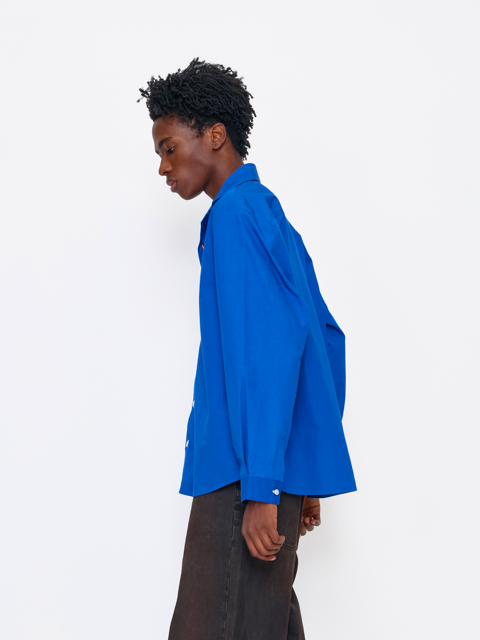 A model shown in profile wearing a bright royal blue button-up shirt and dark brown trousers, standing against a clean white studio background.