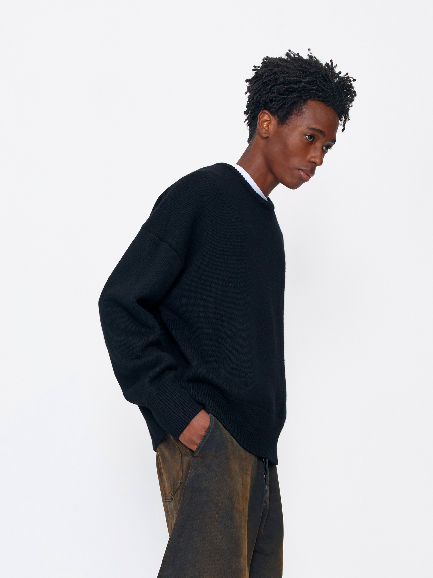A man wearing a black knit crewneck sweater over a white undershirt and brown pants, photographed against a clean white studio background.