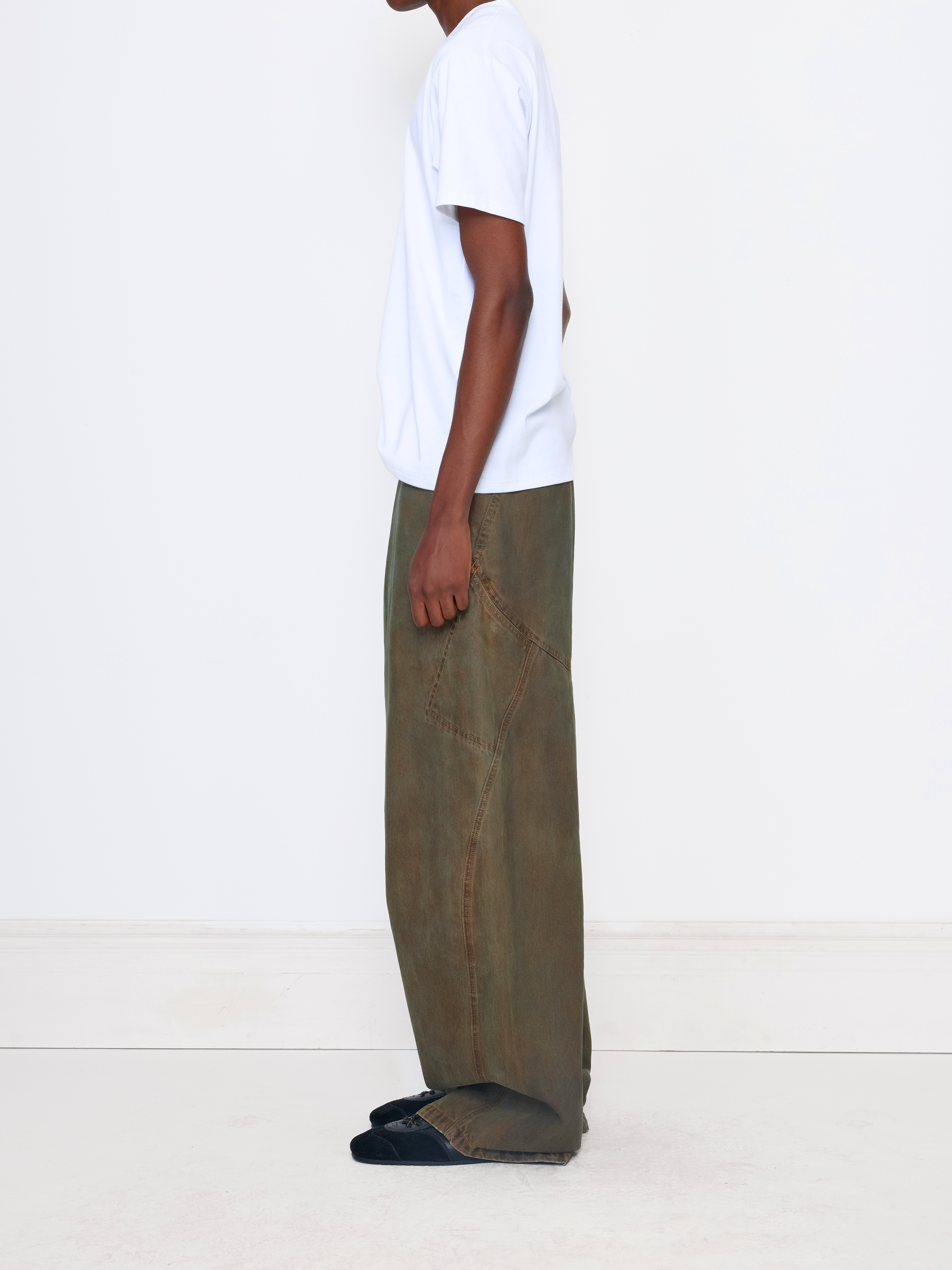A side-profile view of a model wearing wide-leg, relaxed-fit olive green trousers, a white t-shirt, and dark slip-on shoes against a clean white studio background.