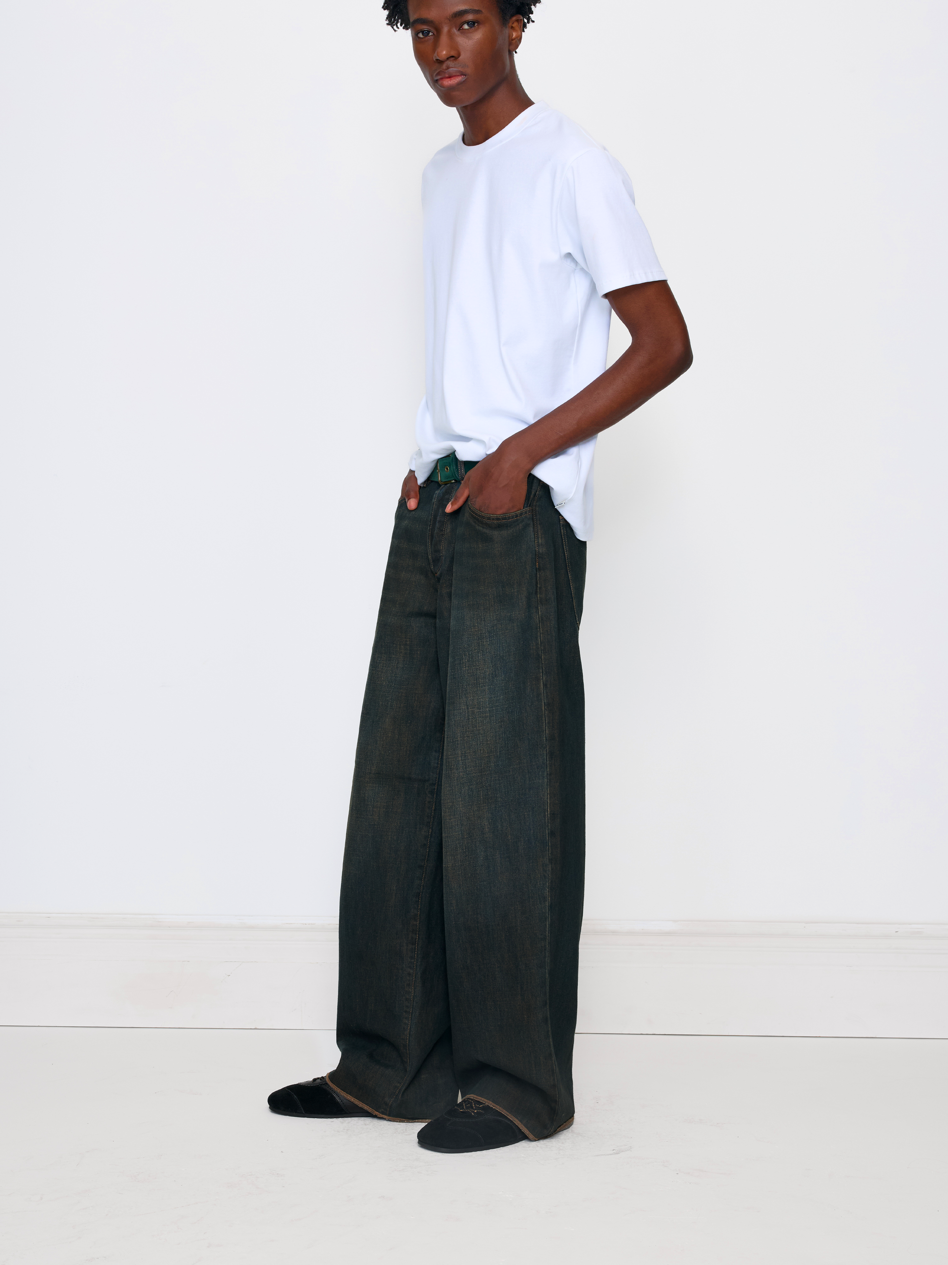 A model stands in a studio wearing a white crew-neck t-shirt, dark wide-leg denim jeans, and black slip-on shoes against a plain white background.