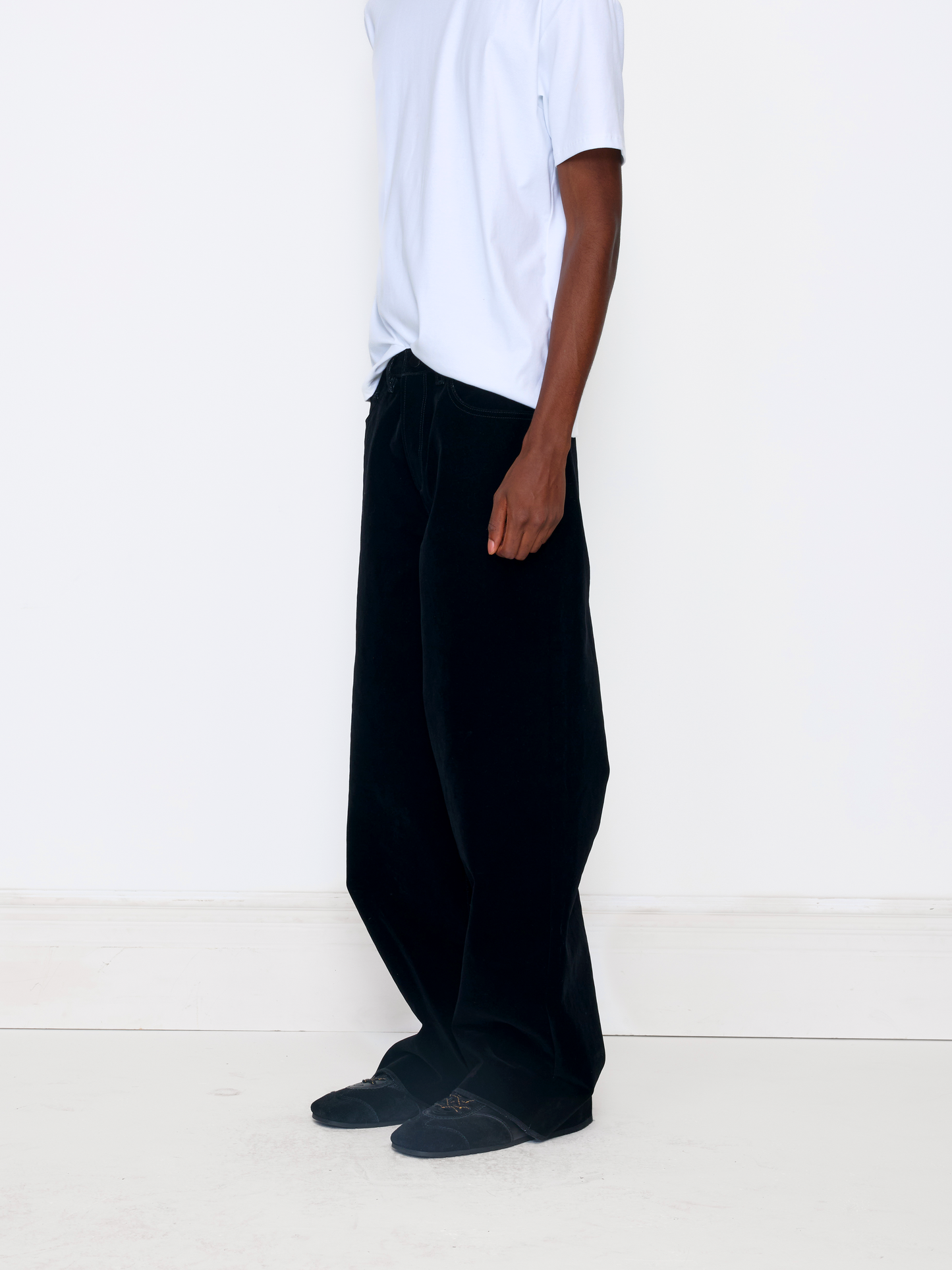 A model wearing black wide-leg corduroy trousers, styled with a white t-shirt and dark slip-on shoes, standing against a plain white wall.